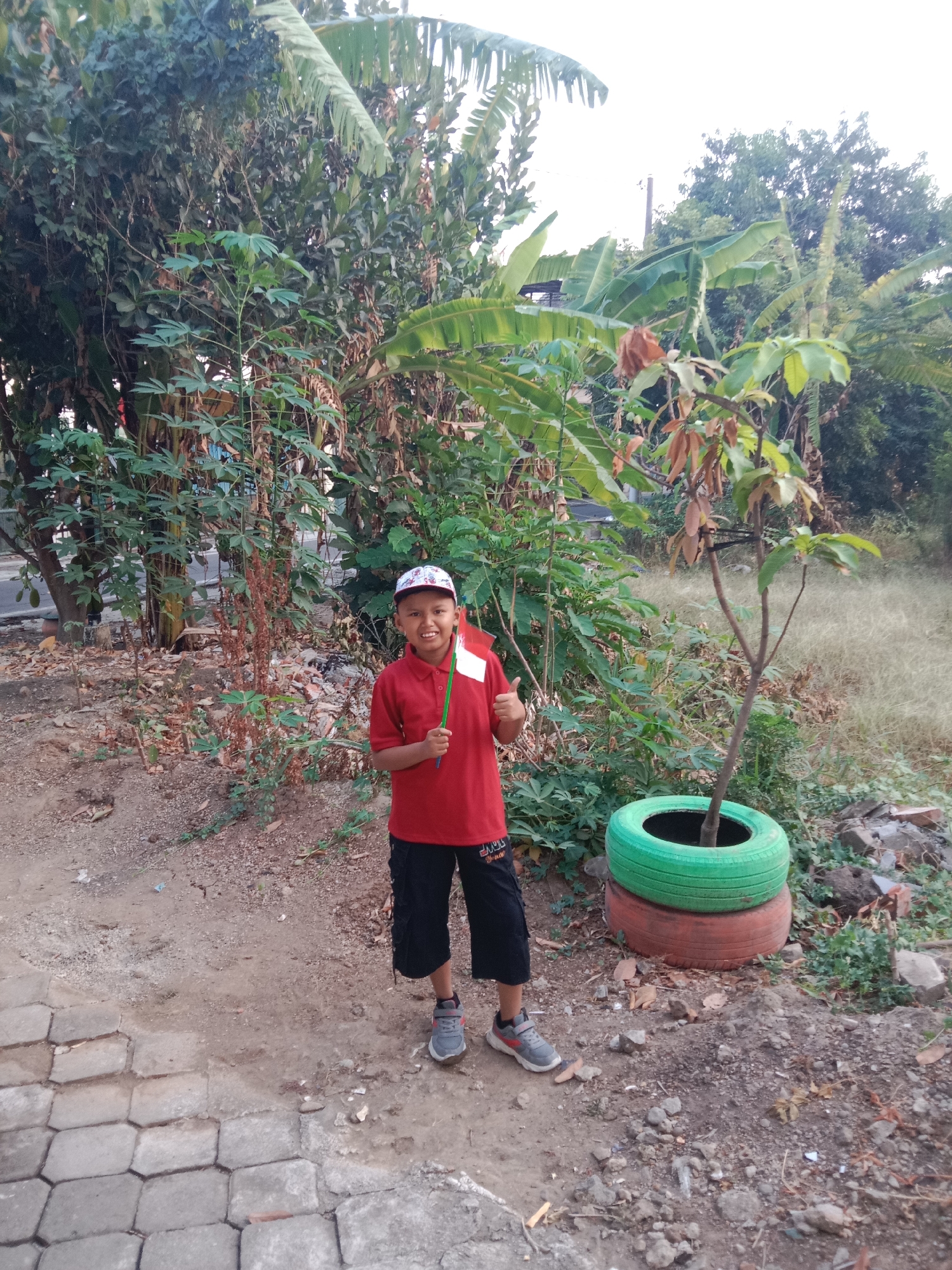 A young Indonesian boy wearing a red polo shirt, black shorts, a baseball cap, and gray sneakers stands outside, holding a small Indonesian flag. He is smiling and looking at the camera. The background consists of dense vegetation, including banana trees and other plants. To the right of the boy, a young tree is planted in two stacked, brightly colored tires, one green and one orange. The ground is a mix of dirt and gravel, with a paved walkway on the left side of the frame. The overall impression is of a rural or semi-rural setting in Madiun, Indonesia, possibly near a residential area. The lighting suggests it is daytime, with soft sunlight filtering through the foliage.