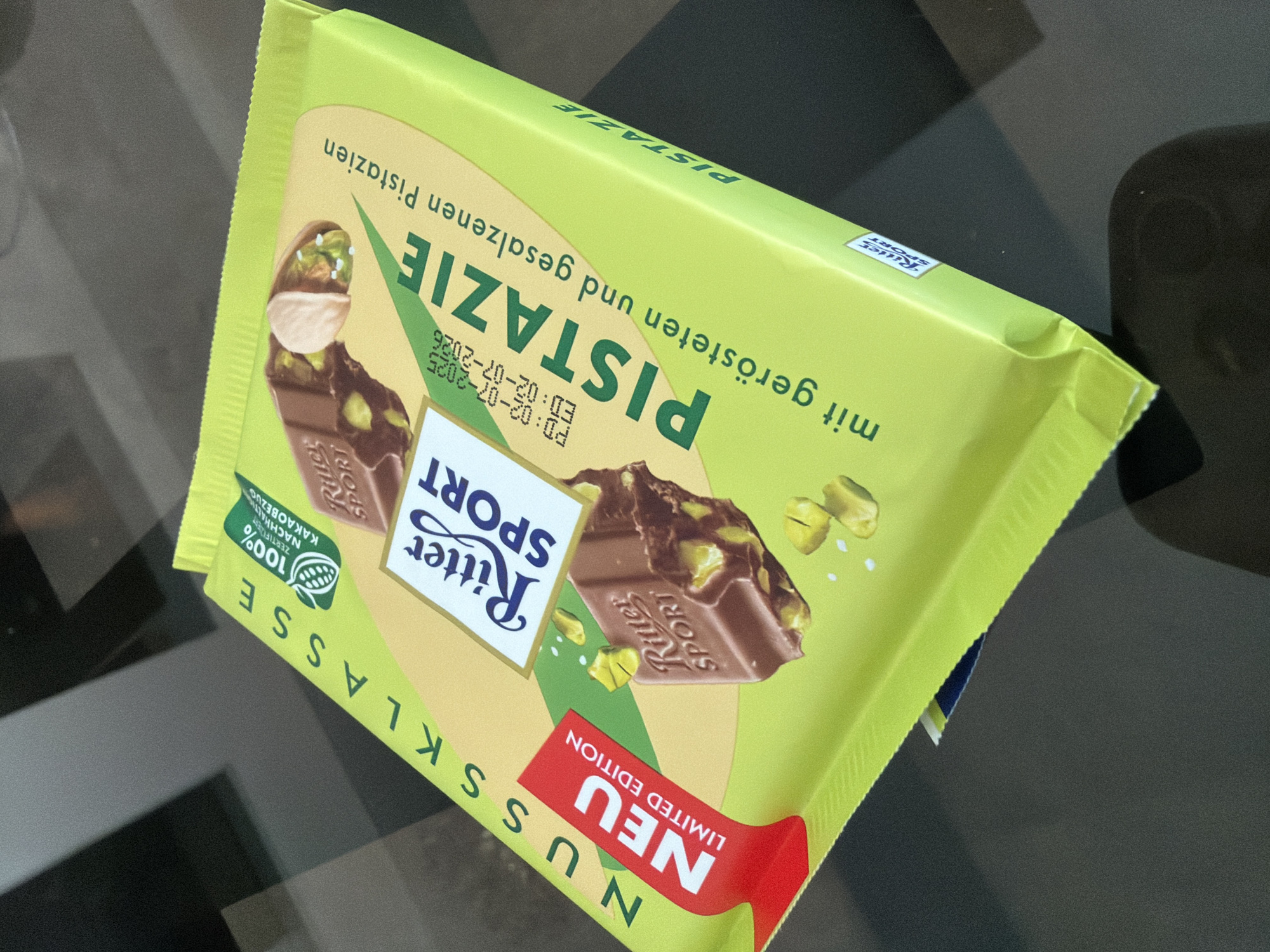 The image shows a Ritter Sport chocolate bar with pistachios, presented on a dark, reflective surface. The packaging is predominantly bright green with yellow accents. The chocolate bar itself is a square, with the Ritter Sport logo visible. The text on the packaging is in German, indicating "Pistazie" (pistachio) and "mit gerösteten und gesalzenen Pistazien" (with roasted and salted pistachios). There is also a red "NEU LIMITED EDITION" sticker. The lighting suggests an indoor environment with overhead light sources creating reflections on the surface. There are no people or animals visible in the image. The background is out of focus, showing dark and light areas that appear to be the reflection of an indoor space.