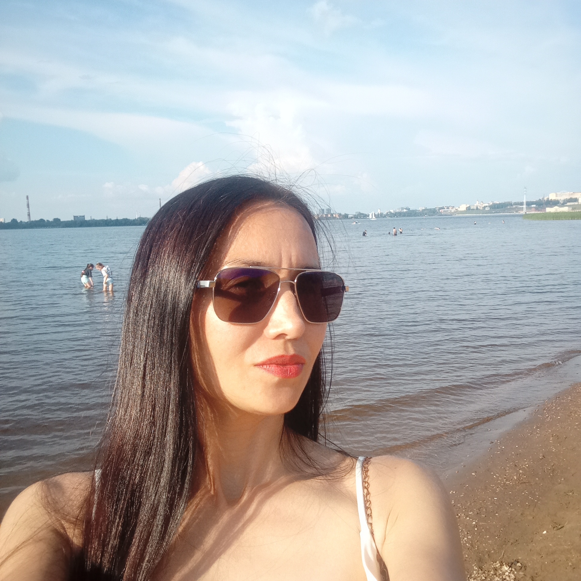 A woman with long, dark hair and sunglasses is taking a selfie at the beach in Izhevsk, Russia. She is wearing a white bikini top with a gold chain strap. In the background, several people are wading in the water, and a cityscape with industrial buildings is visible on the horizon. The sky is blue with scattered clouds.