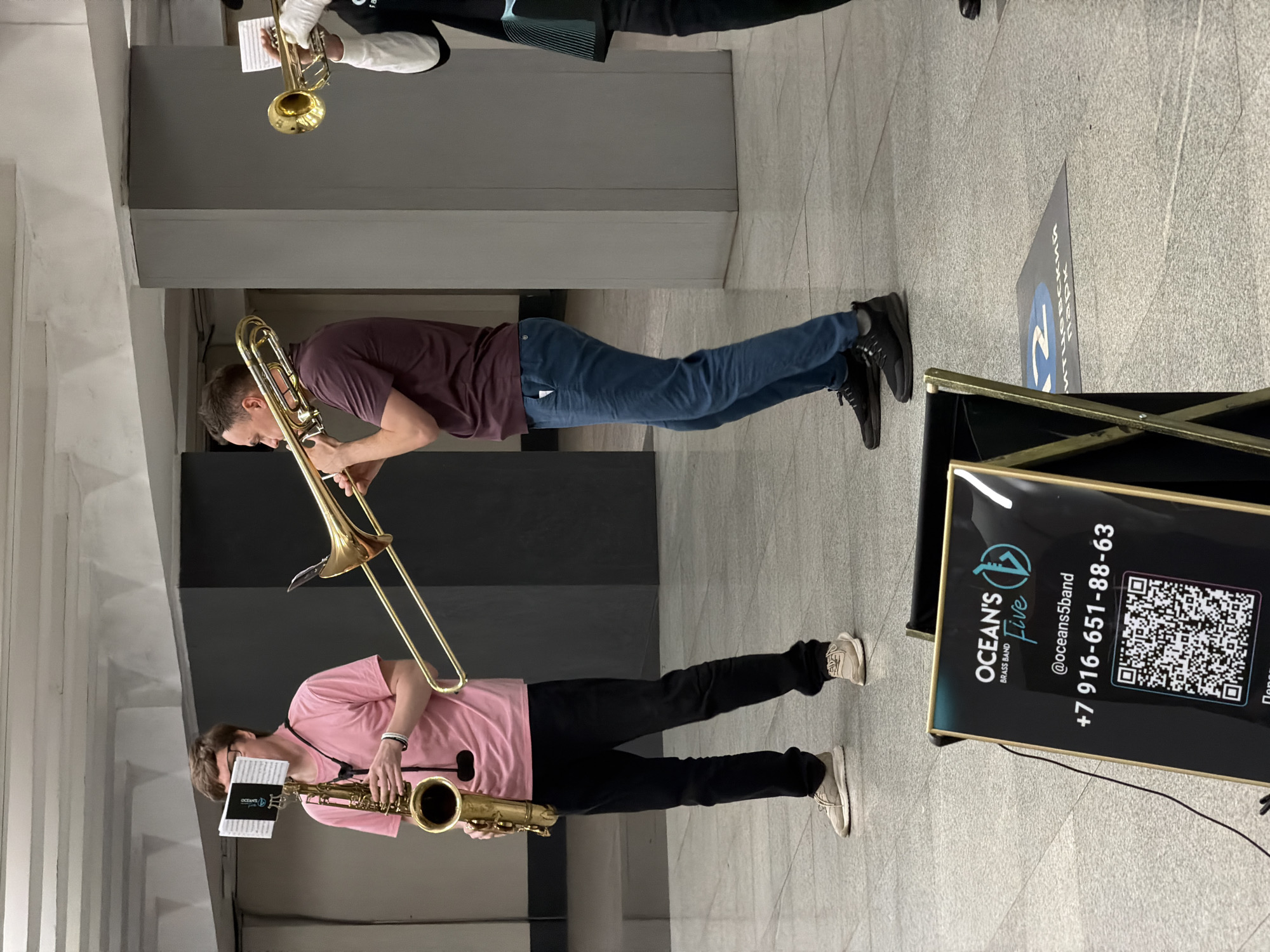 In a Moscow metro station, three musicians perform. A man in a pink t-shirt plays a saxophone, with sheet music attached to its bell. To his right, a man in a burgundy t-shirt plays a trombone. Another musician, partially visible on the right, plays a trumpet. In the foreground, a black stand displays information for "OCEAN'S FIVE BRASS BAND", including a QR code and a phone number. The floor is tiled in a light gray granite pattern, and the ceiling features exposed concrete beams. A blue circle with the number "12" and Cyrillic text is visible on the floor in the background.