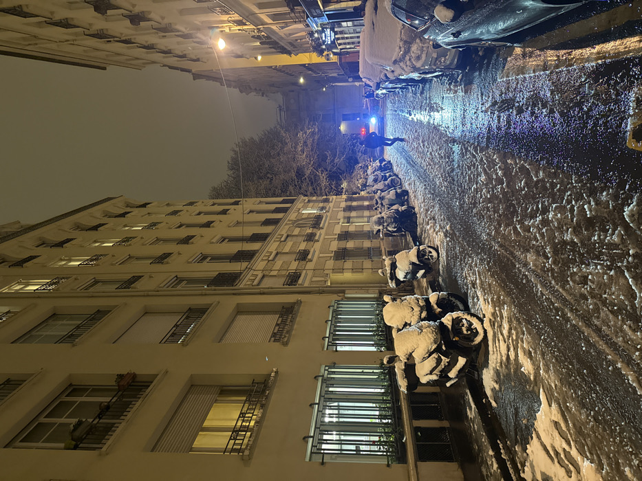 A nighttime urban scene shows a snow-covered street lined with parked cars and motorcycles. A person walks on the street towards a vehicle with flashing blue lights. Buildings line one side of the street, their windows illuminated. The road surface is wet and icy, reflecting the light from street lamps and the vehicle's lights. Snow is visible on the vehicles and motorcycles. The sky is overcast.