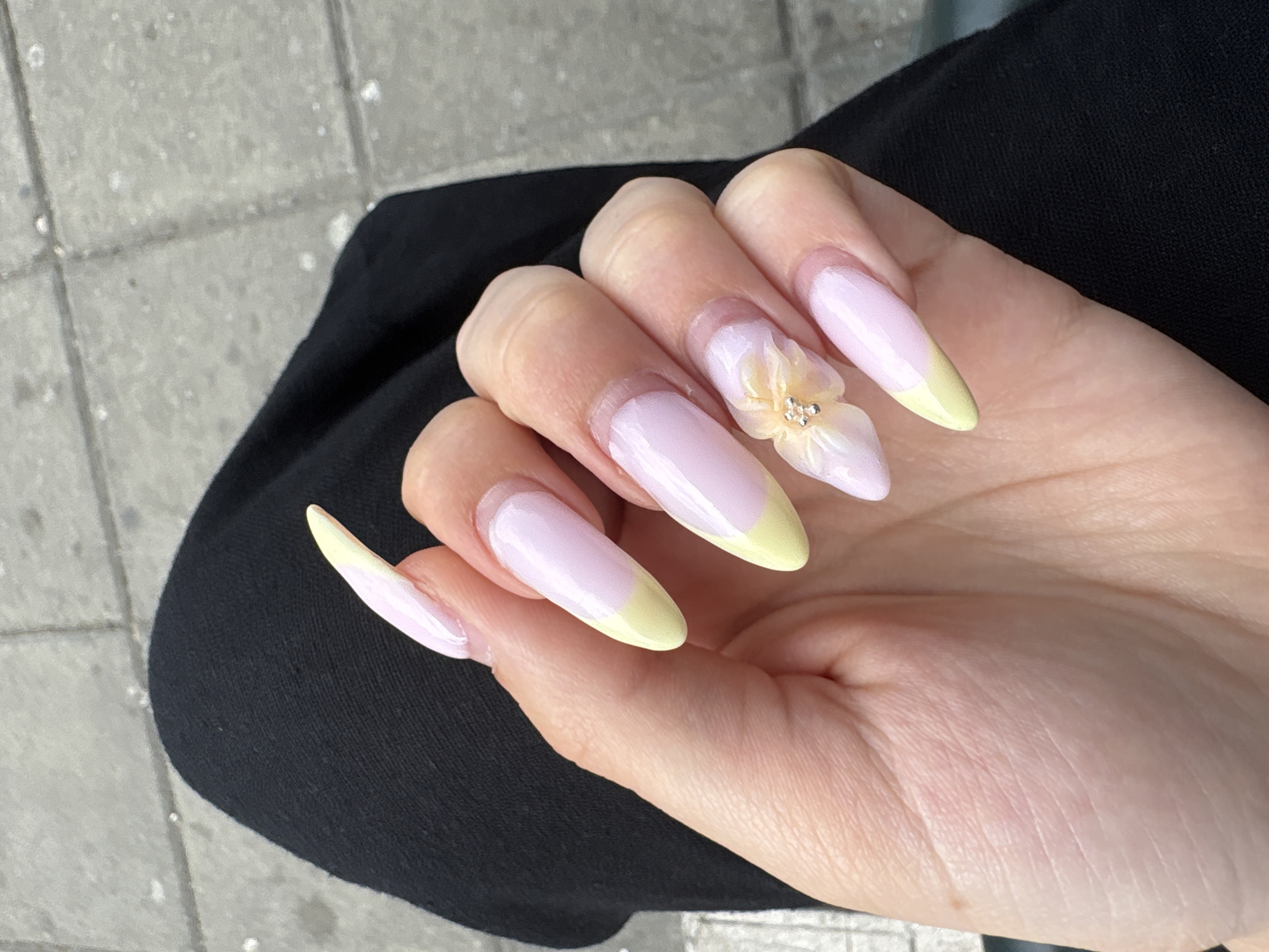 A person's hand with long, stiletto-shaped nails painted in a pastel pink and pale yellow French tip design is shown. The middle finger nail features a delicate 3D floral embellishment with small silver beads. The hand rests against a black fabric, possibly clothing, and the background appears to be an outdoor urban setting with textured gray pavement made of square tiles. The lighting suggests an overcast day.