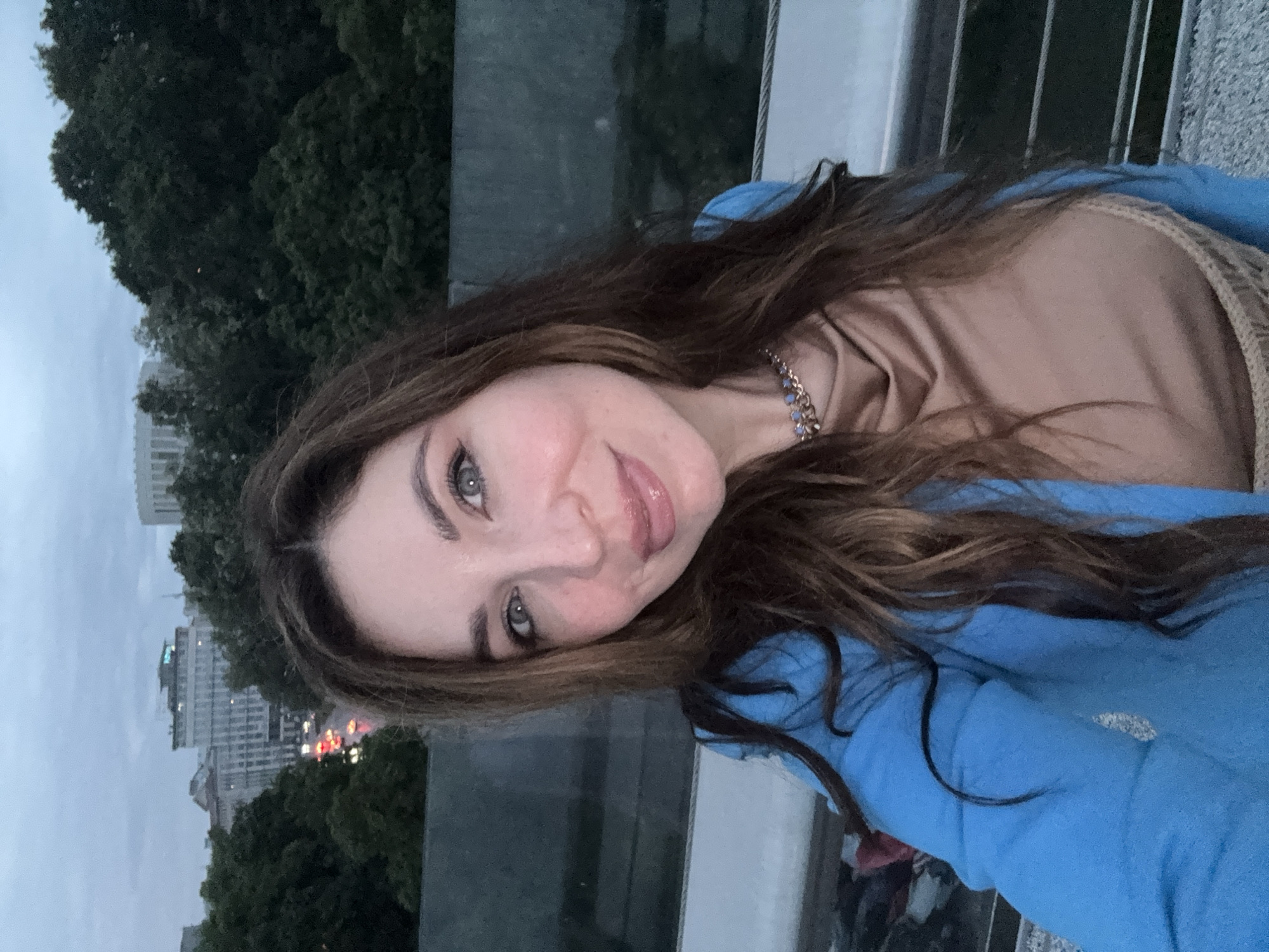 An evening selfie captures a young woman smiling brightly at the camera, positioned against an urban backdrop in Kyiv, Ukraine. She has long, wavy dark brown hair, fair skin with visible freckles, and striking greyish-blue eyes. Her lips are slightly parted in a wide smile, with her tongue just visible. She is wearing a bright blue hoodie or jacket over a beige, high-necked top, and a gold-colored chain necklace with small blue stones is visible around her neck.
The setting appears to be an elevated outdoor location, possibly a bridge or observation deck, indicated by a glass or reflective barrier in the foreground. The background portrays an urban landscape at dusk or early evening, with an overcast grey sky. Dense, dark green trees fill much of the mid-ground, suggesting a park or green area within the city. In the distance, various buildings are silhouetted against the sky, some showing illuminated windows. Red and white lights, likely from city traffic, are faintly visible further back, confirming the late hour and bustling urban environment. The overall mood is friendly and candid. No text is visible in the image.