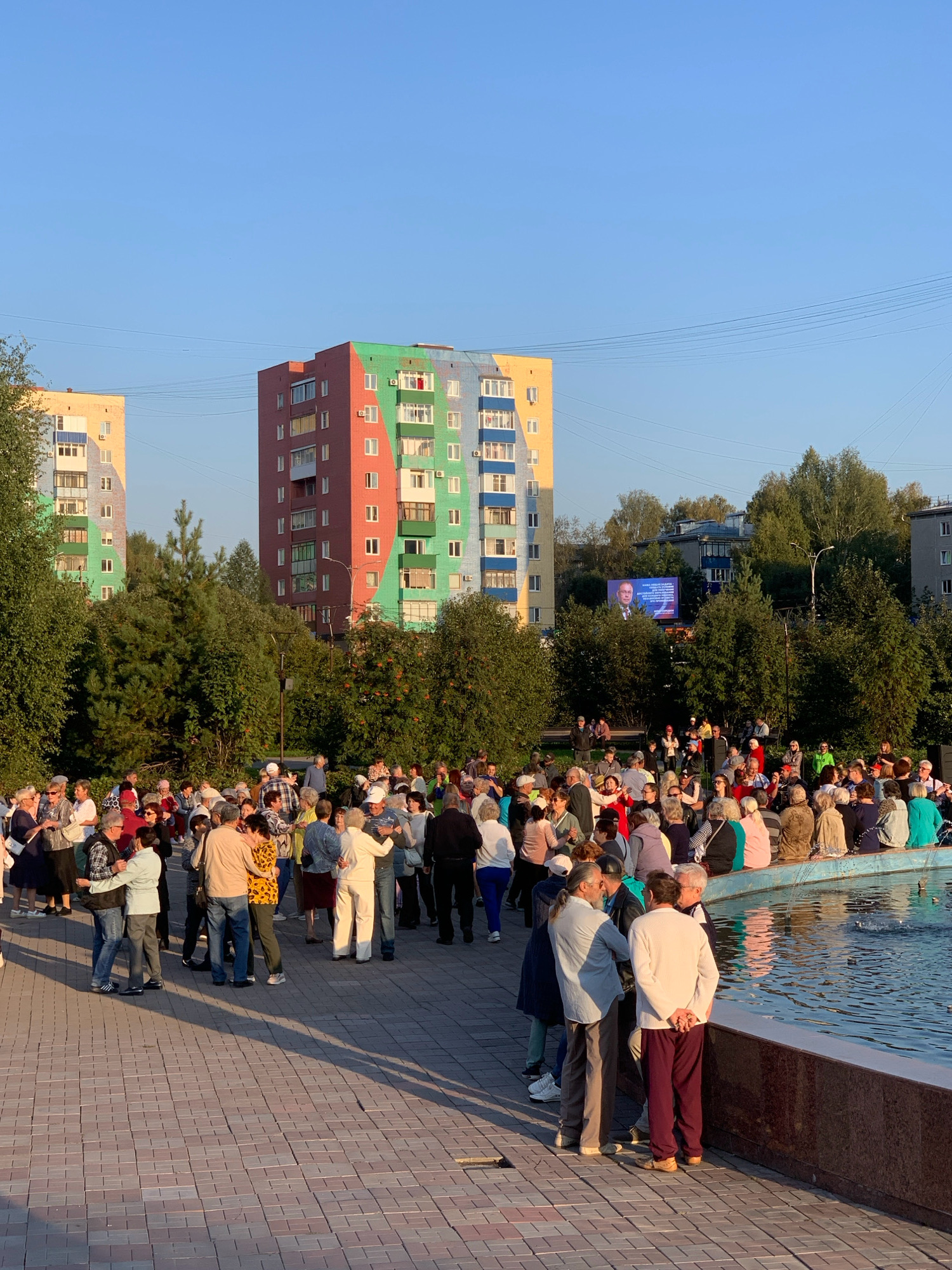 The image depicts an outdoor gathering of people in a paved plaza next to a body of water, possibly a fountain, in Prokopyevsk, Russia. In the background, two apartment buildings are visible, one painted in vibrant multi-colored sections of yellow, green, red, and blue, and the other in a similar multi-colored scheme, predominantly yellow and blue. A large screen displaying a portrait of a man and text is also visible in the background, positioned between the buildings and behind a line of trees. A crowd of people, appearing to be mostly adults and seniors, are congregated in the plaza. Some individuals are dancing in pairs, while others are standing in groups, conversing. The setting sun casts long shadows across the paved ground, indicating it is late afternoon or early evening. The environment appears to be a public urban space, likely a park or square, with trees and urban architecture forming the backdrop.
