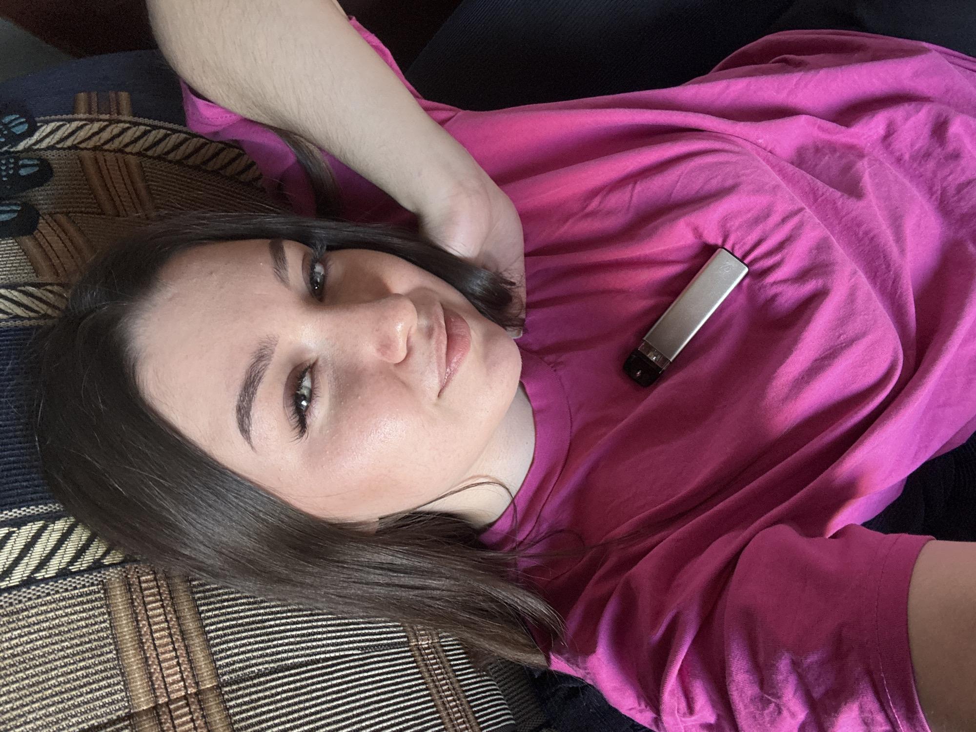 A young woman lies on a striped couch, her head turned towards the camera. She has dark hair and is wearing a bright pink t-shirt. A silver harmonica is clipped to the t-shirt on her chest. The lighting suggests it is daytime, and the scene appears to be indoors, possibly in a home in Krasnokamsk, Russia. The woman's expression is relaxed and she appears to be posing for the photo. There is no visible text in the image.