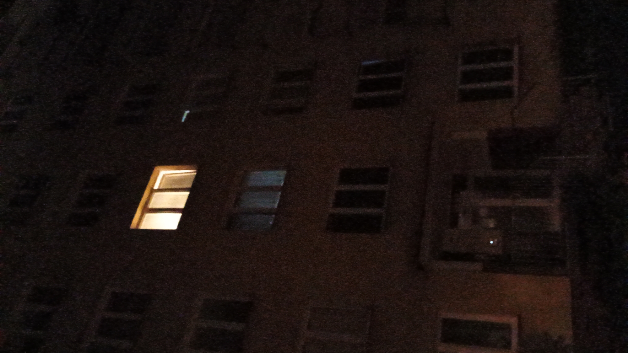 This image is a nighttime, full-frame photograph of the exterior of a multi-story residential building. The building is constructed of a dark, textured material, possibly concrete or brick. Numerous windows are visible on the facade, most of which are dark, indicating they are unlit.
However, one window on the third story from the bottom, near the center-left of the image, is brightly illuminated from within, casting a warm yellow light. This window appears to be divided into three panes. Below this illuminated window, and further to the right on the same floor, there are three other windows that are dimly lit with a cool blue light.
On the ground floor, there is an entrance area with large glass doors, behind which some internal lighting is visible, including a bright circular light. The area in front of the building appears to be a dark, unlit ground-level space, possibly a sidewalk or landscaping.
The overall impression is of a typical apartment building at night, with some residents having their lights on, illuminating their living spaces against the dark facade. The image was taken in Sokolov, Czechia.