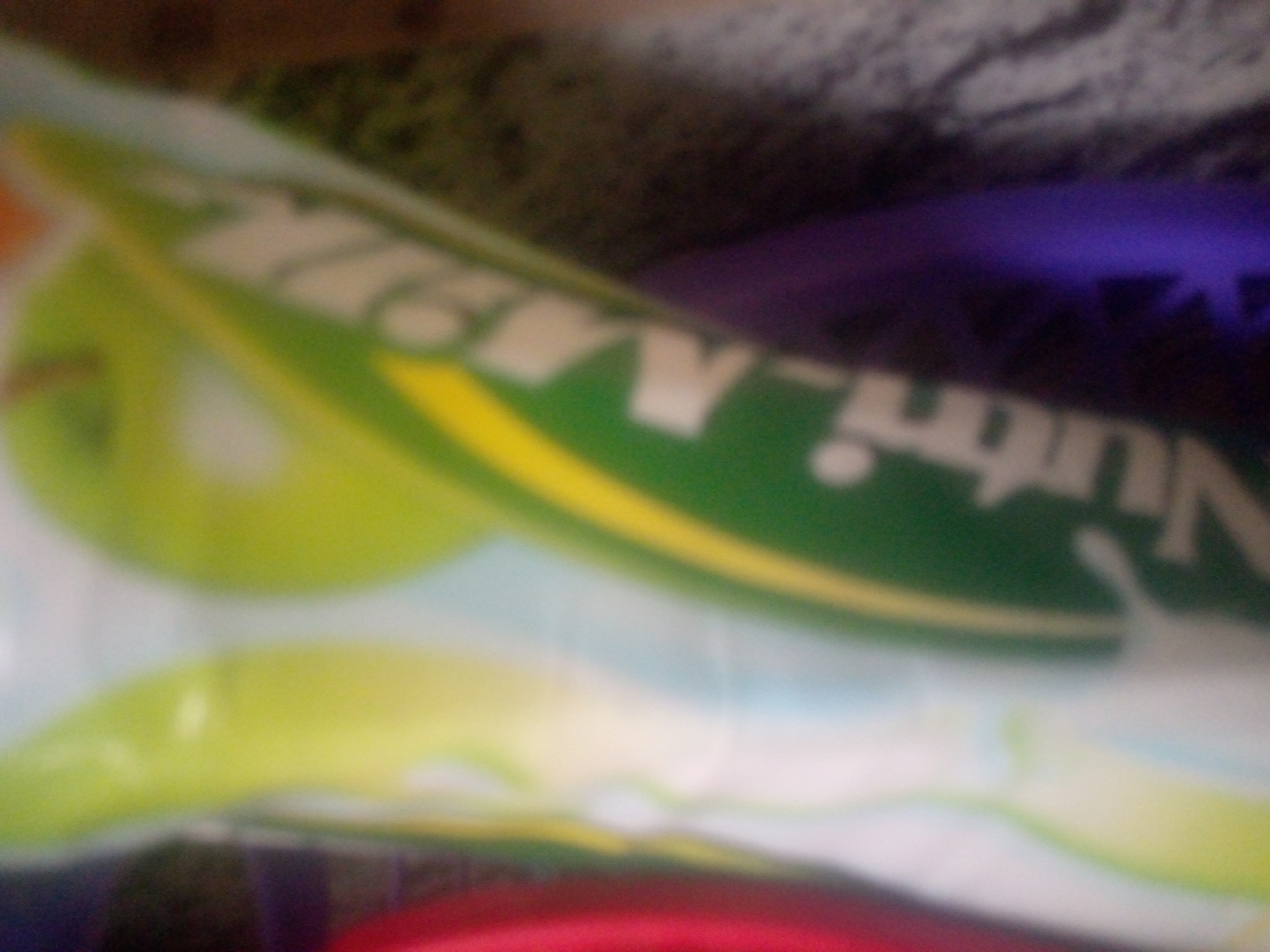 The image is a blurry, close-up photograph of a green and white object with some purple and red elements. The green object has white lettering on it that is partially obscured and difficult to read. There is a yellow stripe running across the green object. The background is dark and out of focus, with a textured surface visible at the top. The overall impression is of a product package or label, possibly for food or a drink, given the colors and stylized lettering. The image is not clear enough to identify any specific location or context beyond the object itself.