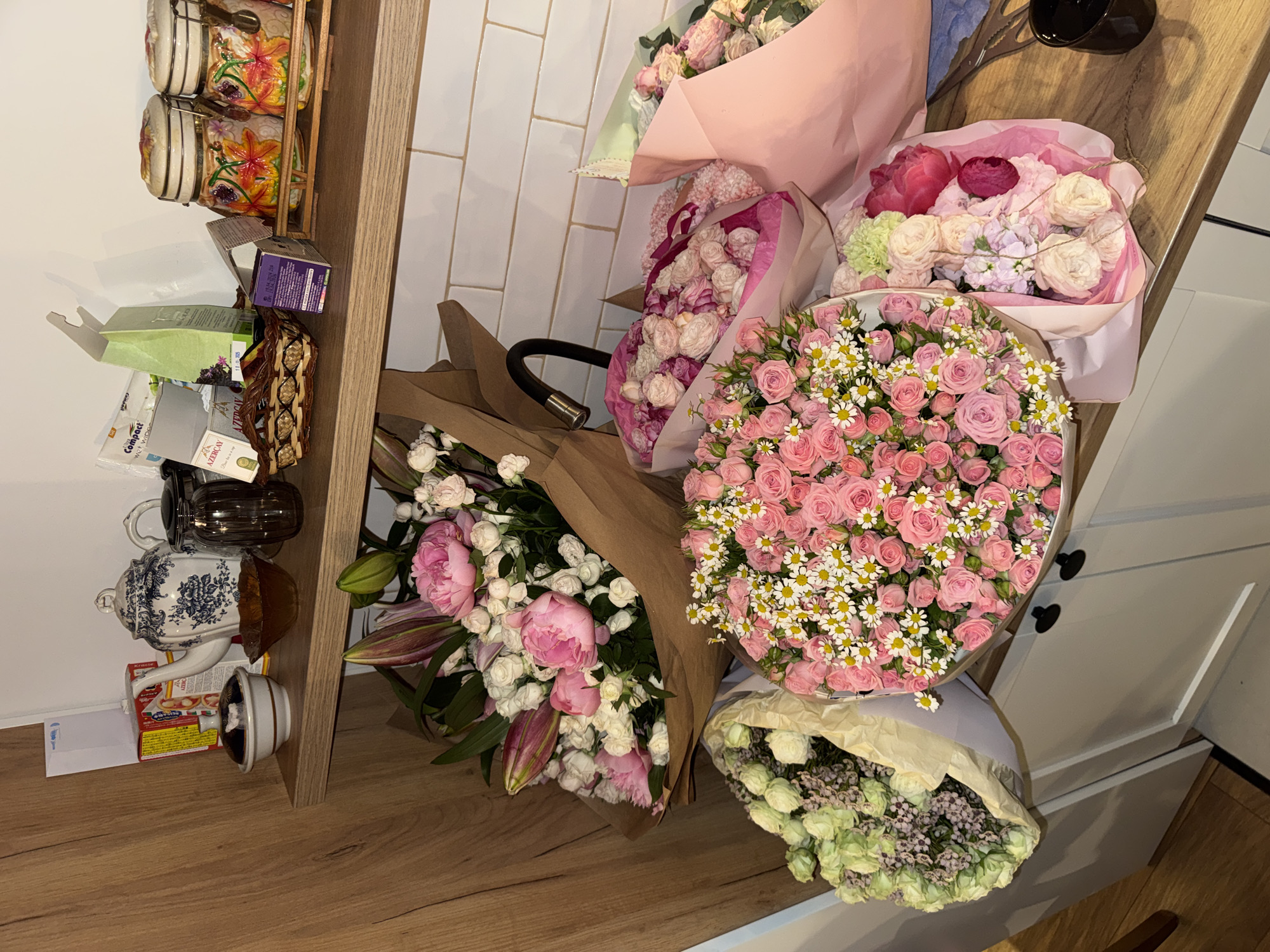 A collection of six bouquets of flowers are arranged on a wooden surface next to a wooden shelf in what appears to be a room. The bouquets are of various colors and styles, including pink roses with white daisies, pink roses, pink and white lilies, white roses and purple flowers, pink and white roses and purple flowers, and a bouquet with a mix of colors including pink, white, and green. The bouquets are wrapped in various materials, including brown paper, pink paper, and light-colored paper. The wooden shelf has various items on it, including decorative jars, boxes, and a teapot. The room has white tiled walls and white cabinets. The location is Unknown, Unknown.
