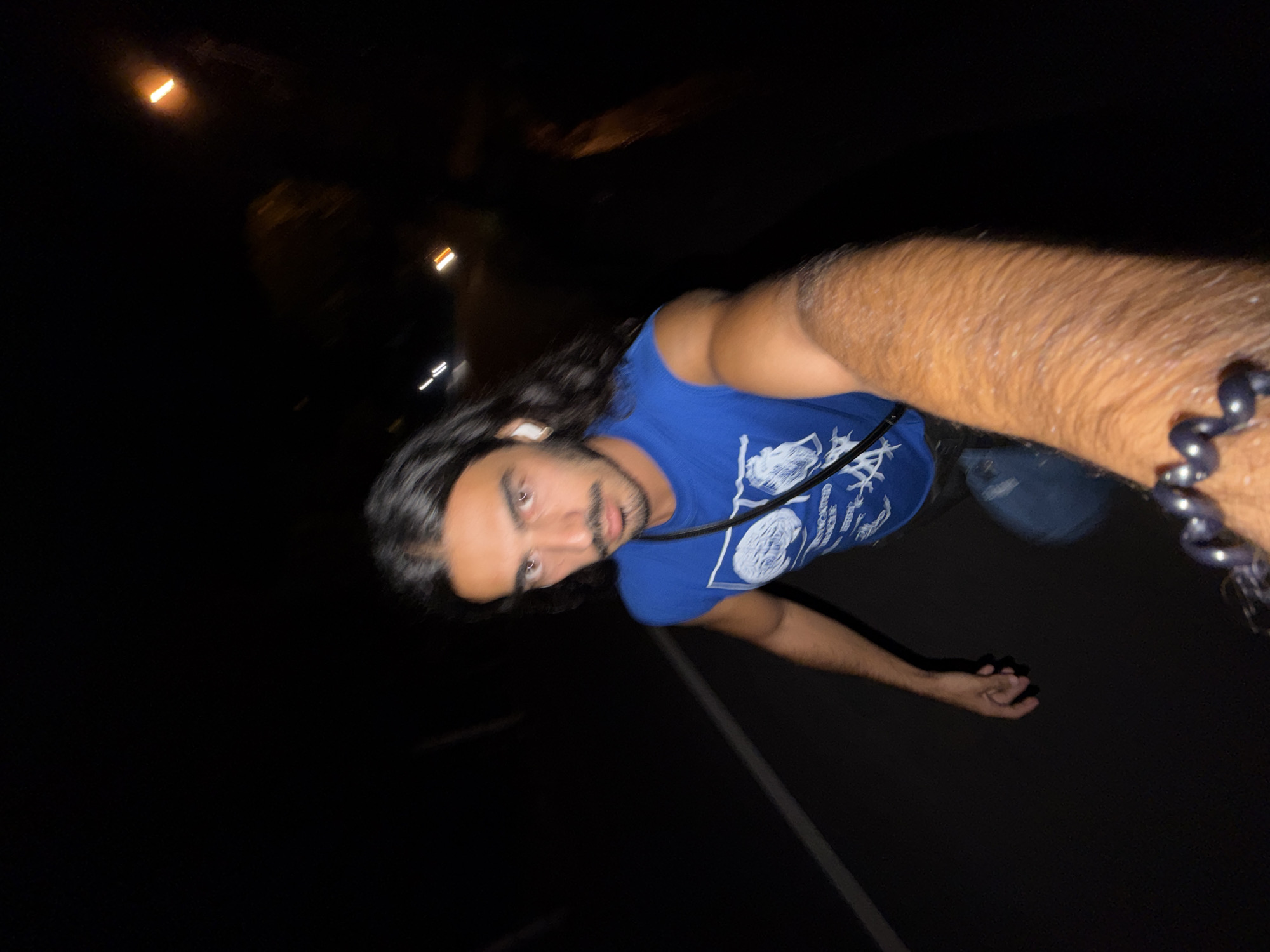A male individual is seen in a selfie taken at night in an outdoor environment, likely in North Kuta, Indonesia. The subject has long dark hair, dark eyes, and a beard. He is wearing a blue tank top and a beaded bracelet. The background is dark with blurred streetlights, indicating an urban setting. The subject appears to be on a road or pavement. The photograph is slightly blurred, suggesting motion or low light conditions.