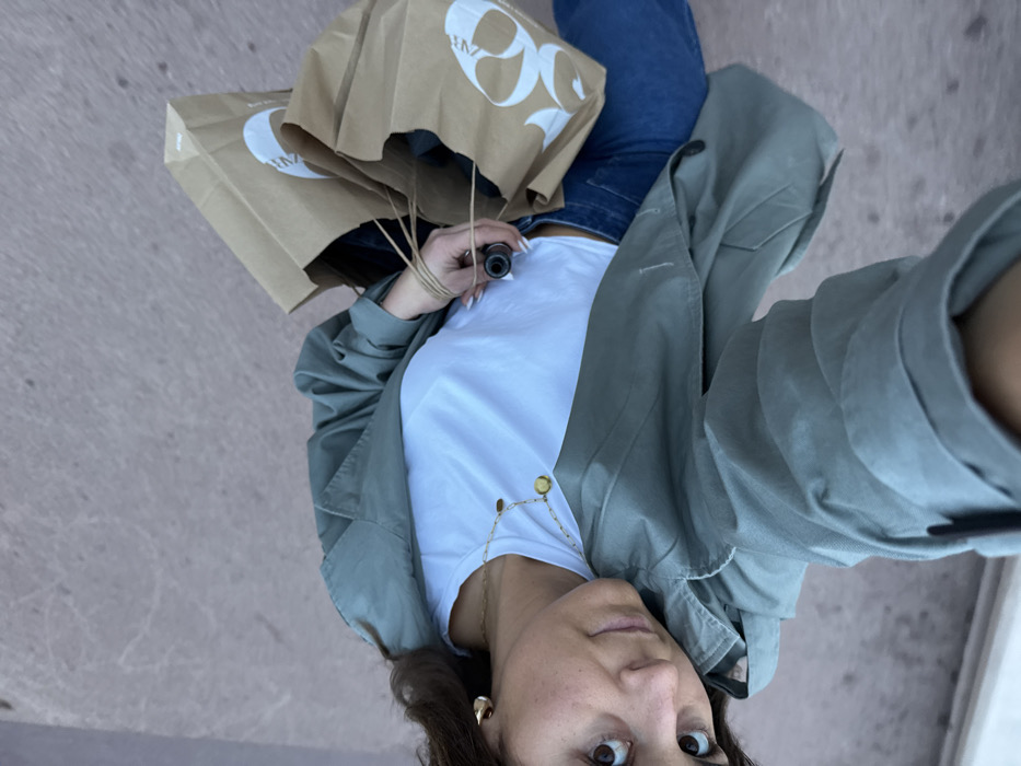 A low-angle selfie captures a person from the waist up, standing on a light grey paved surface. The subject, a woman with brown hair and dark, wide-set eyes, looks directly into the camera with a neutral expression. She is dressed in a casual outfit consisting of a white t-shirt, blue jeans, and a light sage green trench coat or jacket. A long, delicate gold-toned necklace with small circular pendants hangs around her neck, and she wears a matching gold earring.
In her left hand, she holds the string handles of two brown paper shopping bags, both clearly displaying the white "ZARA" logo. Between the fingers of the same hand, she holds a small, black cylindrical object, resembling a vape pen or e-cigarette. The background is a soft, out-of-focus grey, indicating an outdoor setting like a sidewalk or street. The lighting suggests it is daytime. The image conveys a casual moment, possibly taken during a shopping trip in a city, with the provided context suggesting Cannes, France.
