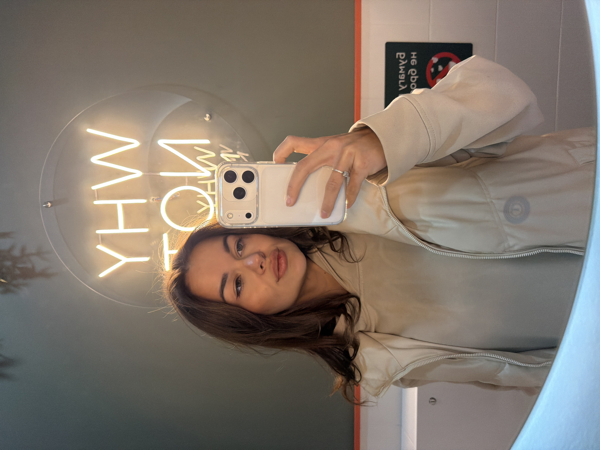 A woman is taking a selfie in a bathroom. She is holding a smartphone, an iPhone, with a clear case in her right hand. Her hair is dark brown and medium length. She is wearing a light beige jacket and a ring on her ring finger. Behind her, a round neon sign spells out "WHY" in reverse. There is a sign indicating "No Smoking." The location is Krasnoyarsk, Russia.