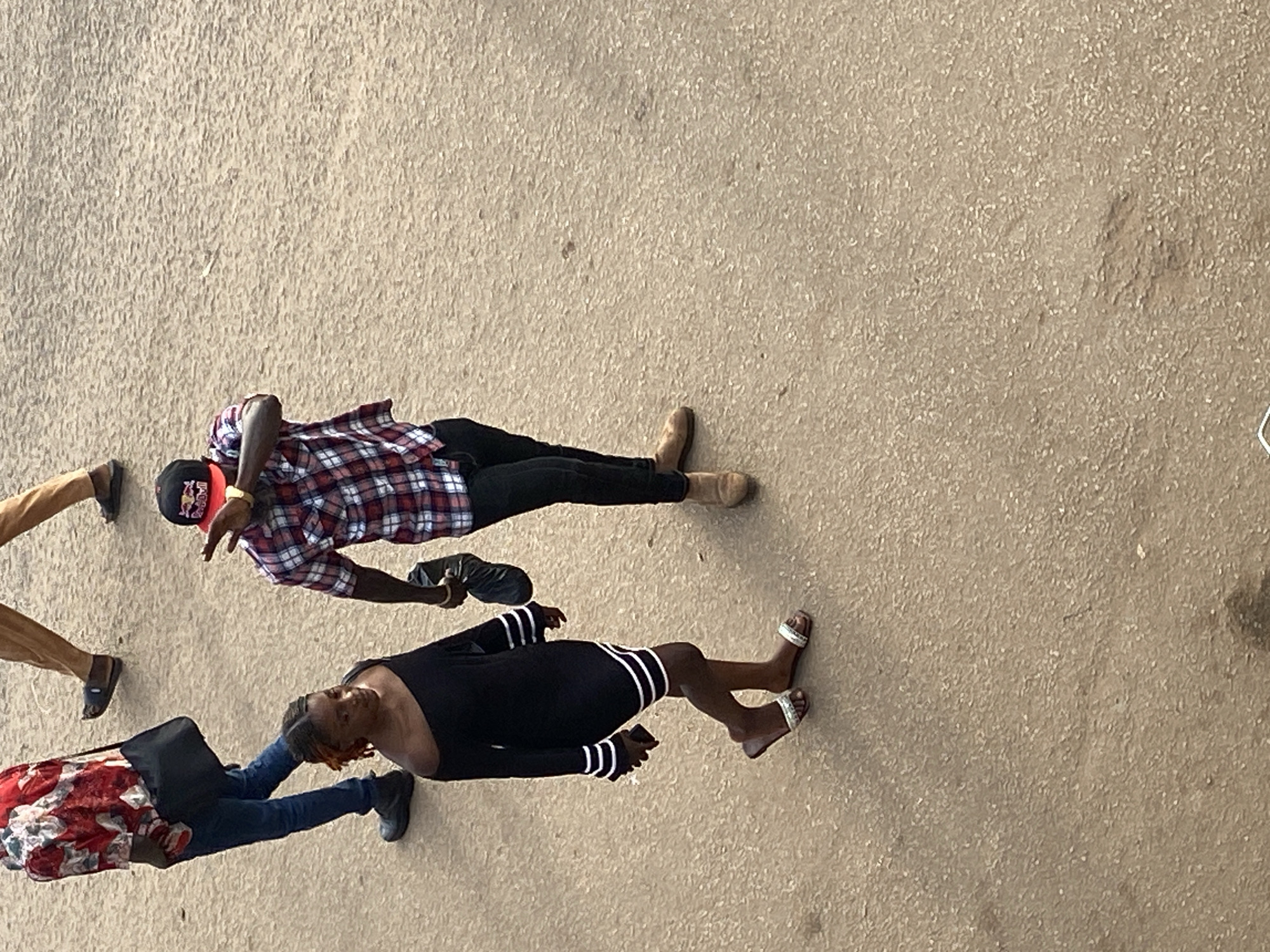 This image captures an overhead view of a street scene in Bukuru, Nigeria, featuring four individuals on a light brown, textured ground surface.
In the upper-left quadrant, a male individual is visible from the waist down, wearing a red and white patterned short-sleeved shirt, blue jeans, and dark footwear. A black messenger bag is slung across his body. He walks toward the right side of the frame.
Adjacent to him, another male individual's lower legs and feet are visible, clad in tan trousers and dark sandals, also moving toward the right.
In the mid-right portion of the frame, a male individual with dark skin walks toward the left. He wears a red, white, and blue plaid short-sleeved button-up shirt, dark trousers, and tan suede-like shoes. A Red Bull baseball cap is worn backward on his head. His right arm is raised with the palm open, seemingly in a gesture or to shield his face. He holds a black plastic bag in his left hand.
In the lower-left quadrant, a female individual with dark skin and reddish-brown hair walks toward the right. She wears a black off-the-shoulder dress featuring two white horizontal stripes on each sleeve and two white horizontal stripes above the hemline. She has silver-colored flat sandals on her feet and holds a dark-colored smartphone in her right hand.
The ground is uniformly light brown, exhibiting a coarse, granular texture consistent with compacted earth or weathered asphalt. The scene is illuminated by natural daylight.