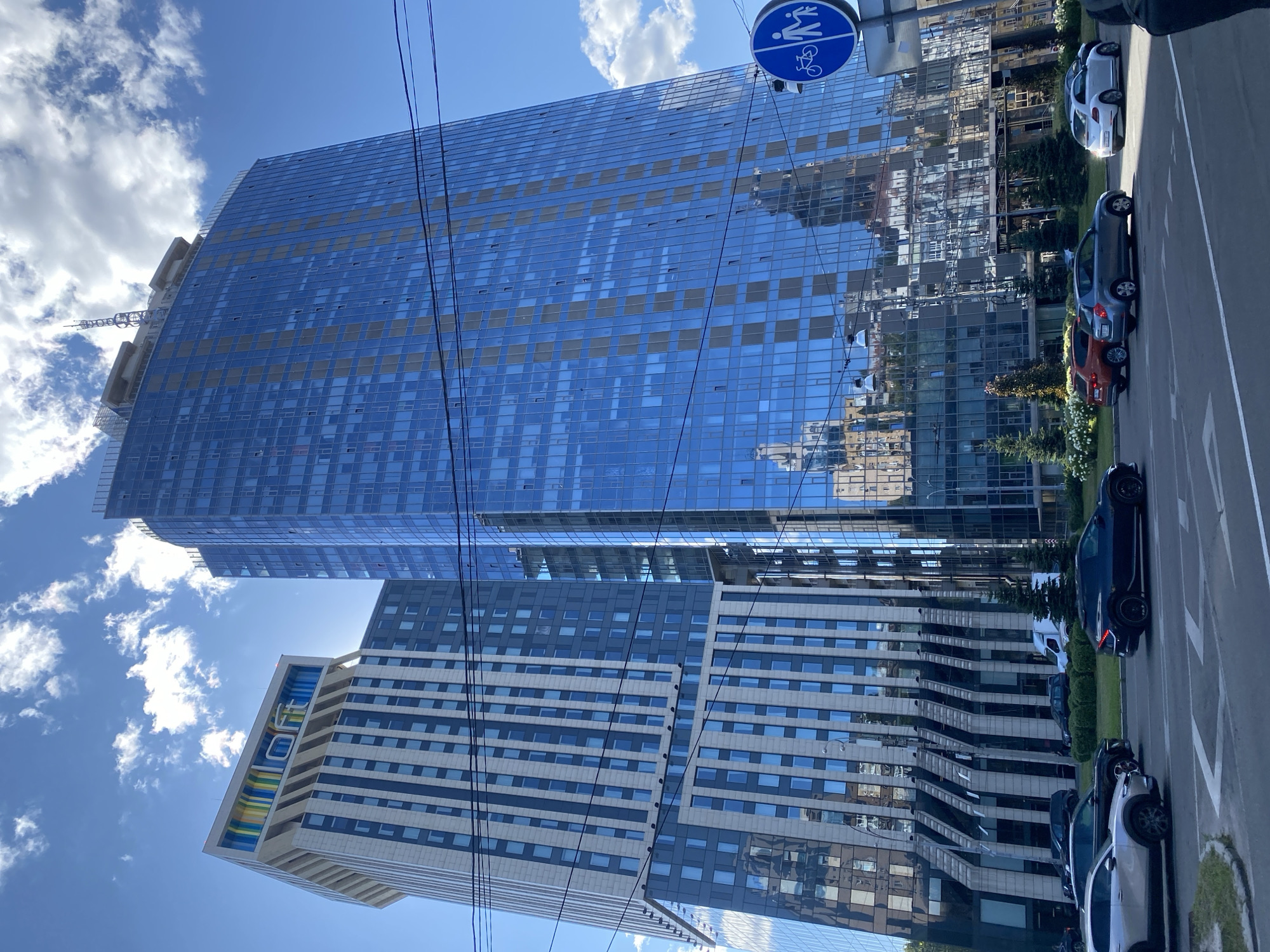 An exterior, eye-level, wide shot captures two modern skyscrapers on a sunny day in an urban environment. The skyscraper on the left has a lighter facade with vertical white and blue-grey stripes, and its peak features a colorful, striped design with the word "LOFT" in white letters. The skyscraper on the right is covered in reflective blue glass, mirroring the sky and surrounding buildings, and has an antenna at its apex. In the foreground, a paved road with white lane markings is visible, with several cars driving on it. These include a silver sedan, a black SUV, an orange hatchback, a grey hatchback, and a white beetle. Green landscaping with trees and shrubs lines the side of the road, behind which the buildings rise. Power lines are strung across the upper portion of the image. A blue circular sign with pictograms of a person walking and a person on a bicycle is mounted on a pole on the right side of the frame. The sky is a clear blue with scattered white clouds.