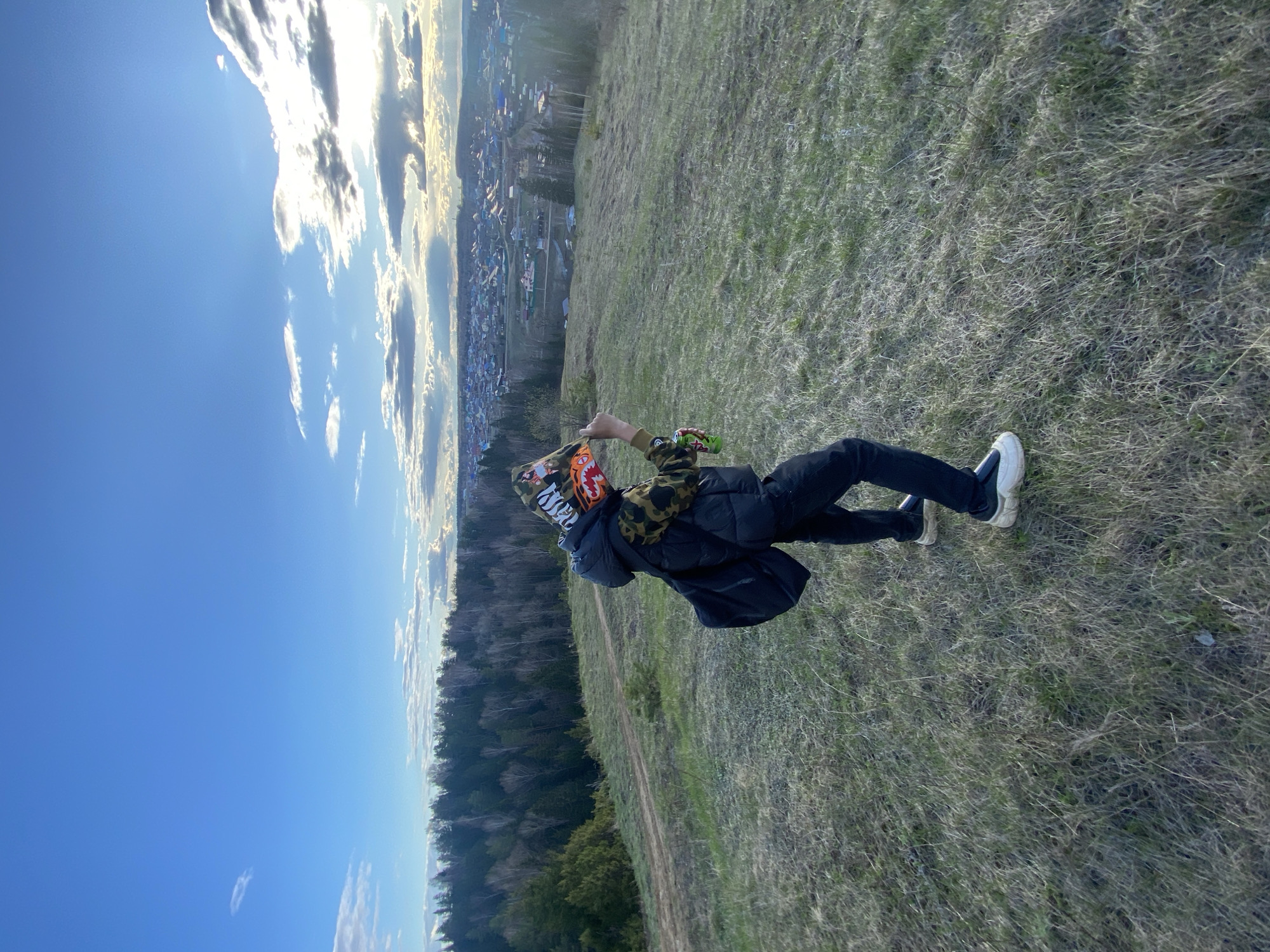 The image captures an outdoor scene featuring a person standing on a grassy hill, viewed from behind, overlooking a distant town and forest. The sky above is bright blue with scattered white and grey clouds, some brilliantly lit, suggesting it is late afternoon or early evening.
The main subject is a person dressed in a dark puffer jacket, black pants, and white sneakers. Their head is covered by a distinctive hoodie or balaclava featuring a camouflage pattern, prominent stylized white text that appears to be "BAPE," and an orange monster-like mouth graphic. The person is holding a green can in their right hand, which is clearly identifiable as a Monster Energy drink by its iconic "M" logo.
The setting is a sprawling natural landscape. The immediate foreground and midground consist of a grassy hillside with patches of both dried and green vegetation. A faint dirt path snakes across the mid-section of the hill. Beyond the hill, a dense forest stretches horizontally, separating the viewer from a populated valley. In the distance, numerous houses of a town or village are visible, nestled among the trees. The overall impression is one of peaceful observation, with the person enjoying the panoramic view from an elevated vantage point.