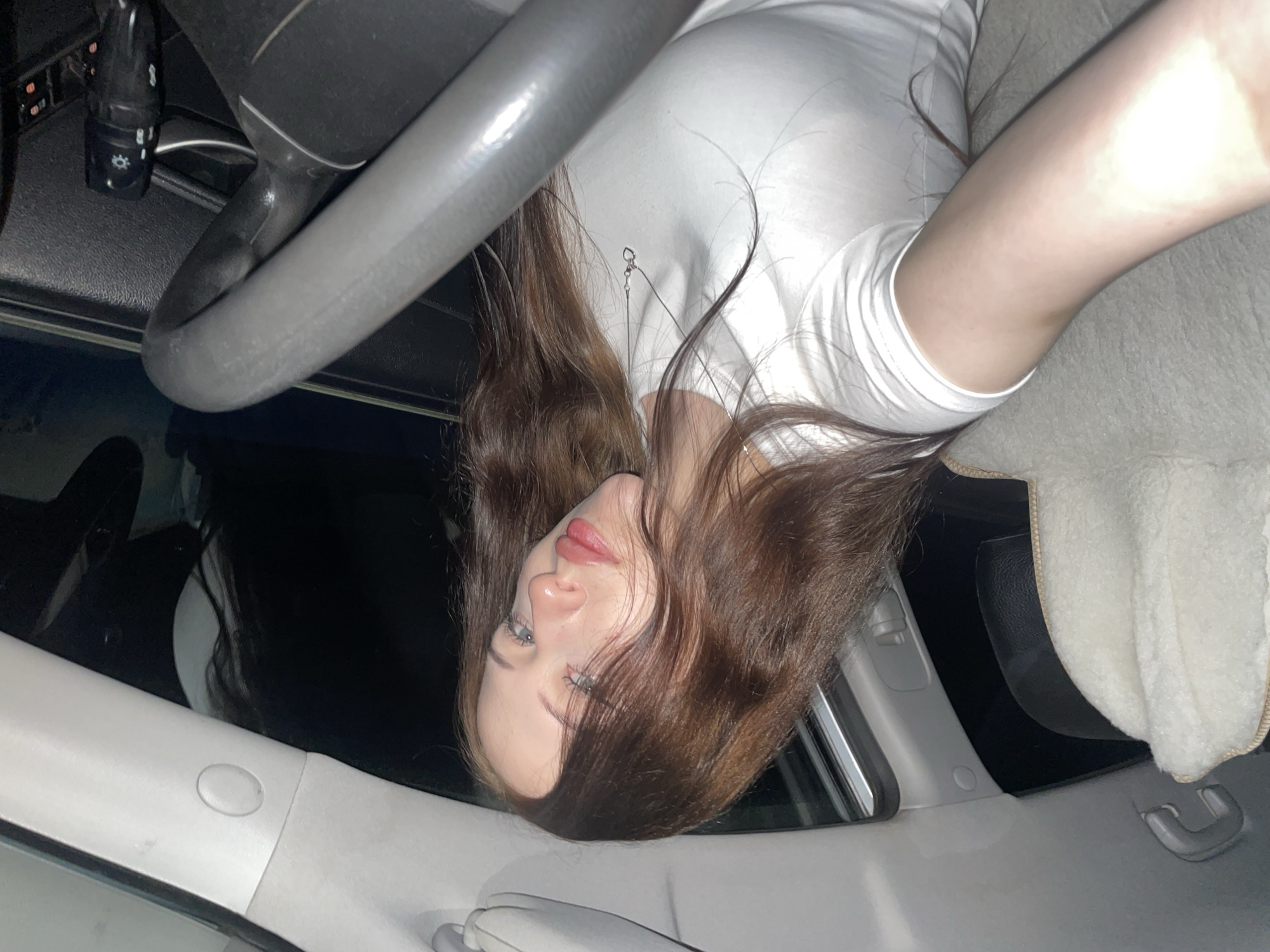 The image shows a young woman inside a car, lying down in the passenger seat. She has long, dark brown hair and is wearing a white t-shirt. The interior of the car appears to be dimly lit, with the steering wheel and dashboard visible. The woman's face is partially obscured by her hair, but her eyes are open and she is looking towards the camera. There is a small, metallic pendant visible on her t-shirt. The overall impression is that of a casual, candid photograph taken at night. The location is stated as Gavrilovka, Russia.