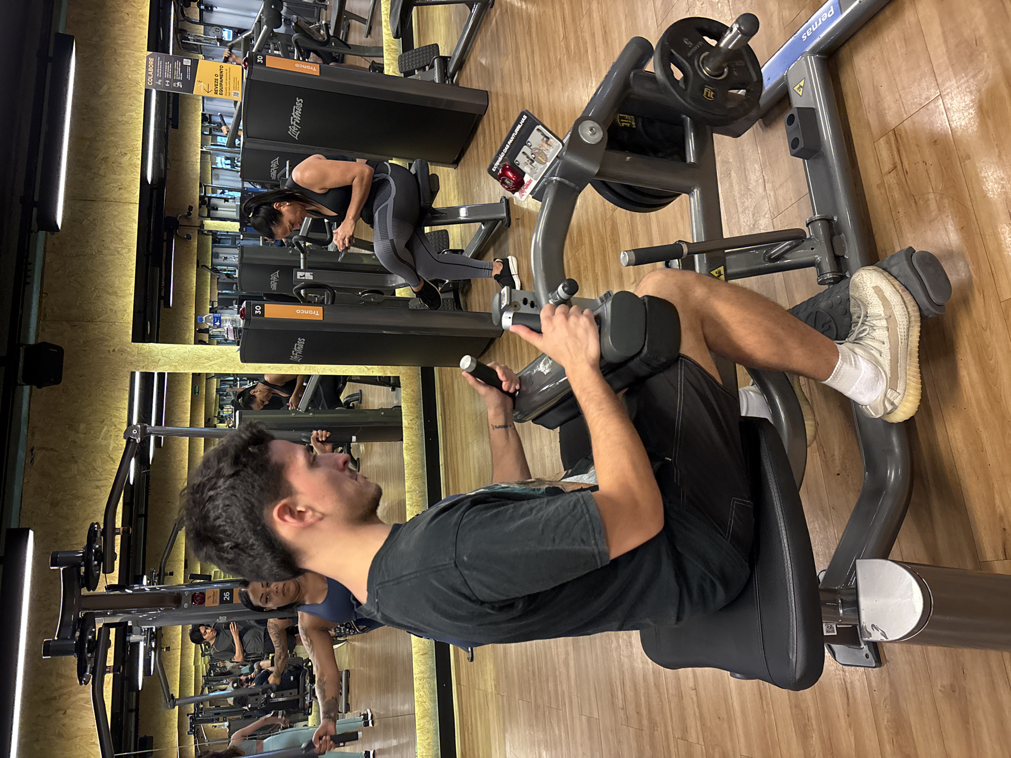 The image depicts the interior of a gym in São Paulo, Brazil. The foreground focuses on a man using a seated calf raise machine with weights. He is wearing a black t-shirt, black shorts, white socks, and light-colored sneakers. Another woman is exercising on a different machine behind him. Multiple other gym-goers and machines are visible in the background. Mirrors on the walls multiply the people visible in the image. Signs in Portuguese are on the machines and walls. The floor is wooden.