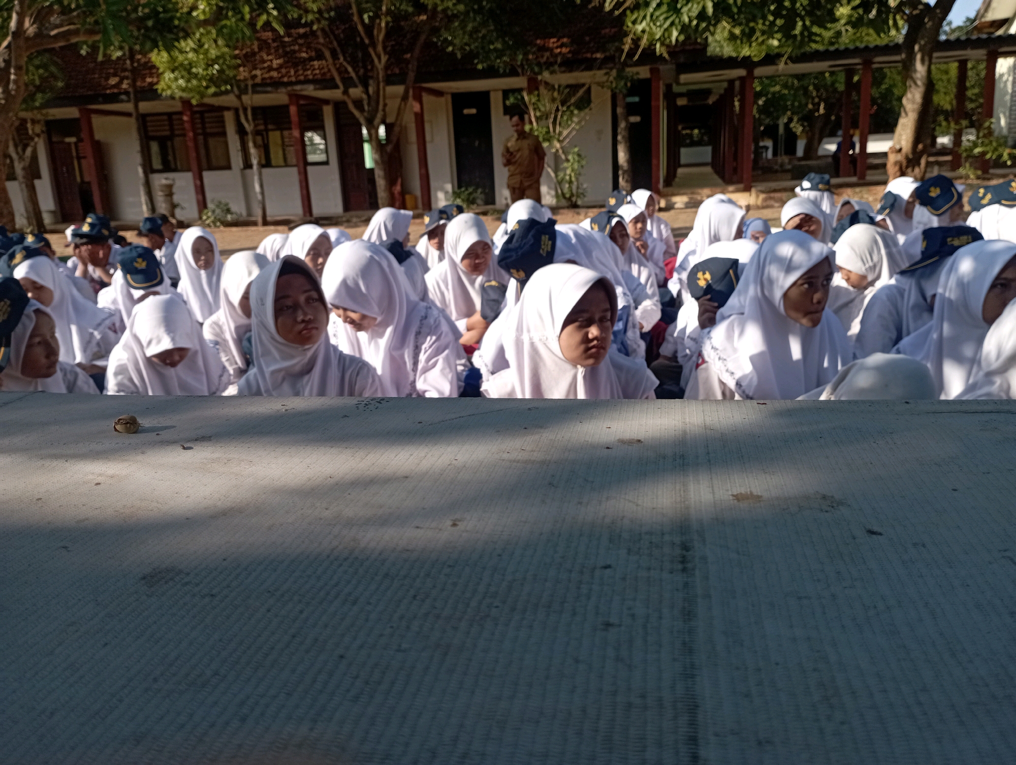 In Banyuurip, Indonesia, a group of young people, likely students, are gathered outdoors in front of a building with a covered walkway. Most are wearing white uniforms with headscarves and some are wearing dark caps with a yellow emblem. They are sitting and appear to be listening or watching something. The foreground features a textured surface, possibly a table or mat, with shadows cast upon it. Trees with green foliage are visible behind the group, suggesting a sunny day. A man in a light brown uniform stands in the background, observing the scene.