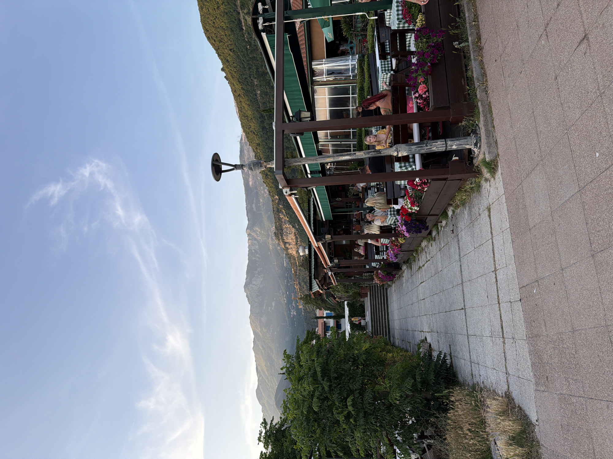 The image captures an outdoor scene in Budva, Montenegro, featuring a partially covered restaurant terrace positioned along a pedestrian walkway. The terrace is lined with wooden railings adorned with flower boxes filled with colorful blooms. Tables with green-and-white checkered tablecloths are occupied by patrons. The background reveals a view of the mountains and a clear blue sky with wispy clouds. A row of streetlights is visible along the terrace. The pedestrian walkway is paved with gray and brown tiles. A dense green tree is visible near the bottom of the image.