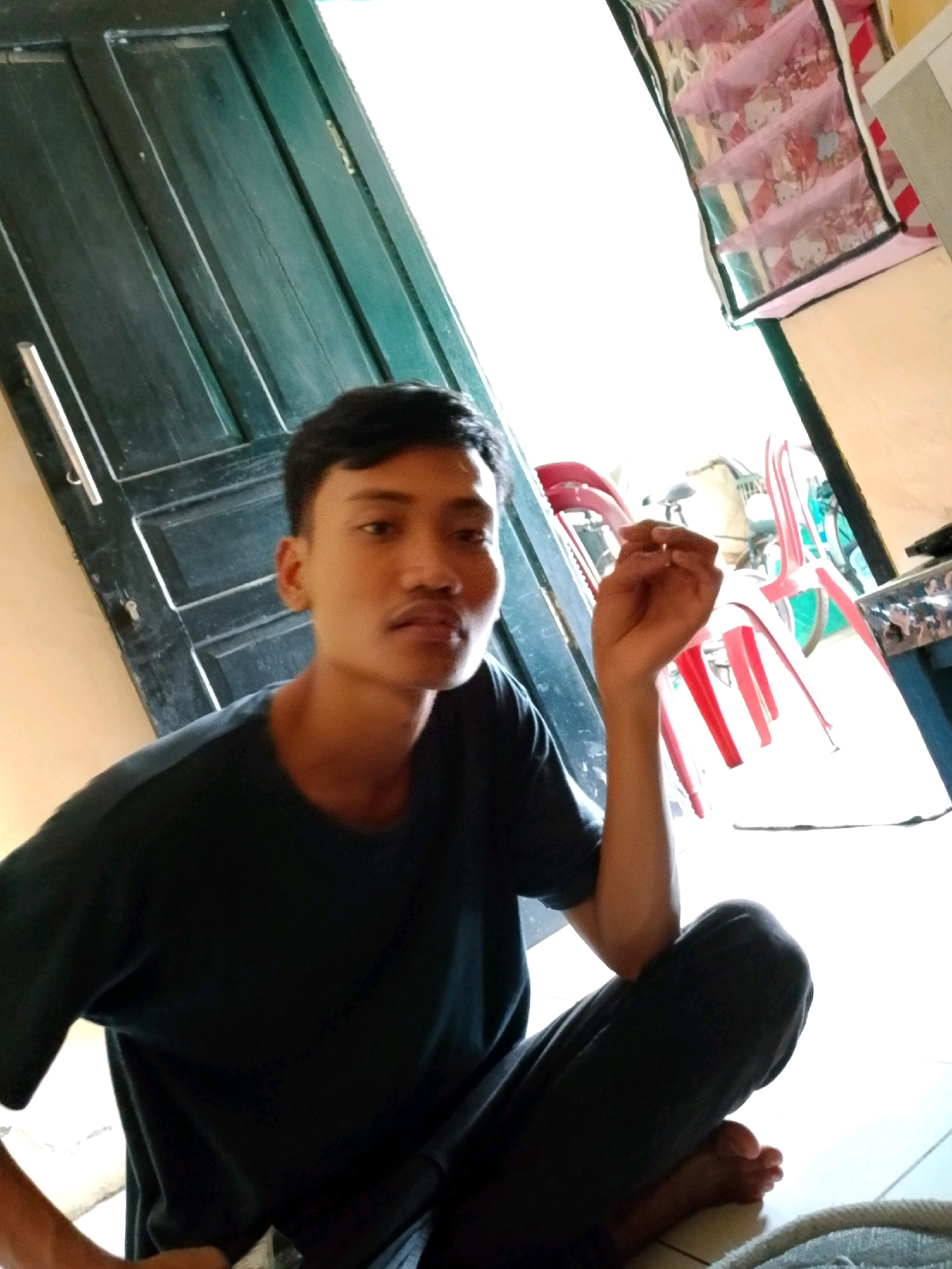 A young man with dark hair and brown eyes is sitting on the floor in front of a dark green wooden door. He is wearing a dark t-shirt and dark pants. His right hand is raised, and he appears to be holding something small and translucent between his fingers. The background is bright and shows several red chairs and what appears to be a shelf with pink fabric dividers. A white appliance is also visible on the right side of the image. The flooring is a light-colored tile. The scene appears to be indoors, possibly in a home or small establishment, located in Tajurhalang, Indonesia.