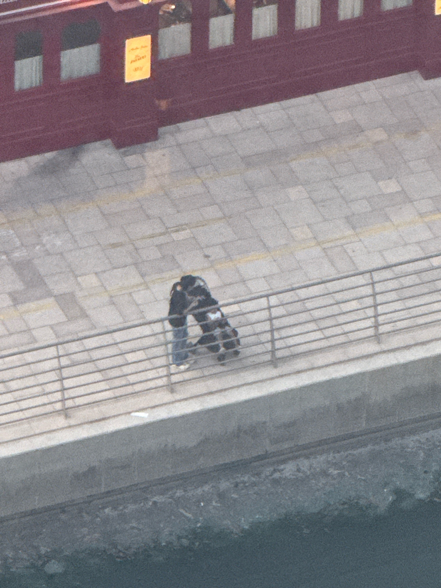 The image shows two individuals embracing on a paved walkway next to a metal railing. The walkway is composed of light-colored rectangular tiles with darker grout lines. A dark red structure with windows is visible in the background, and a sign with yellow text is affixed to its wall. Below the walkway is a concrete structure that appears to be a seawall or embankment, and below that is dark water. The overall scene suggests an outdoor urban environment.