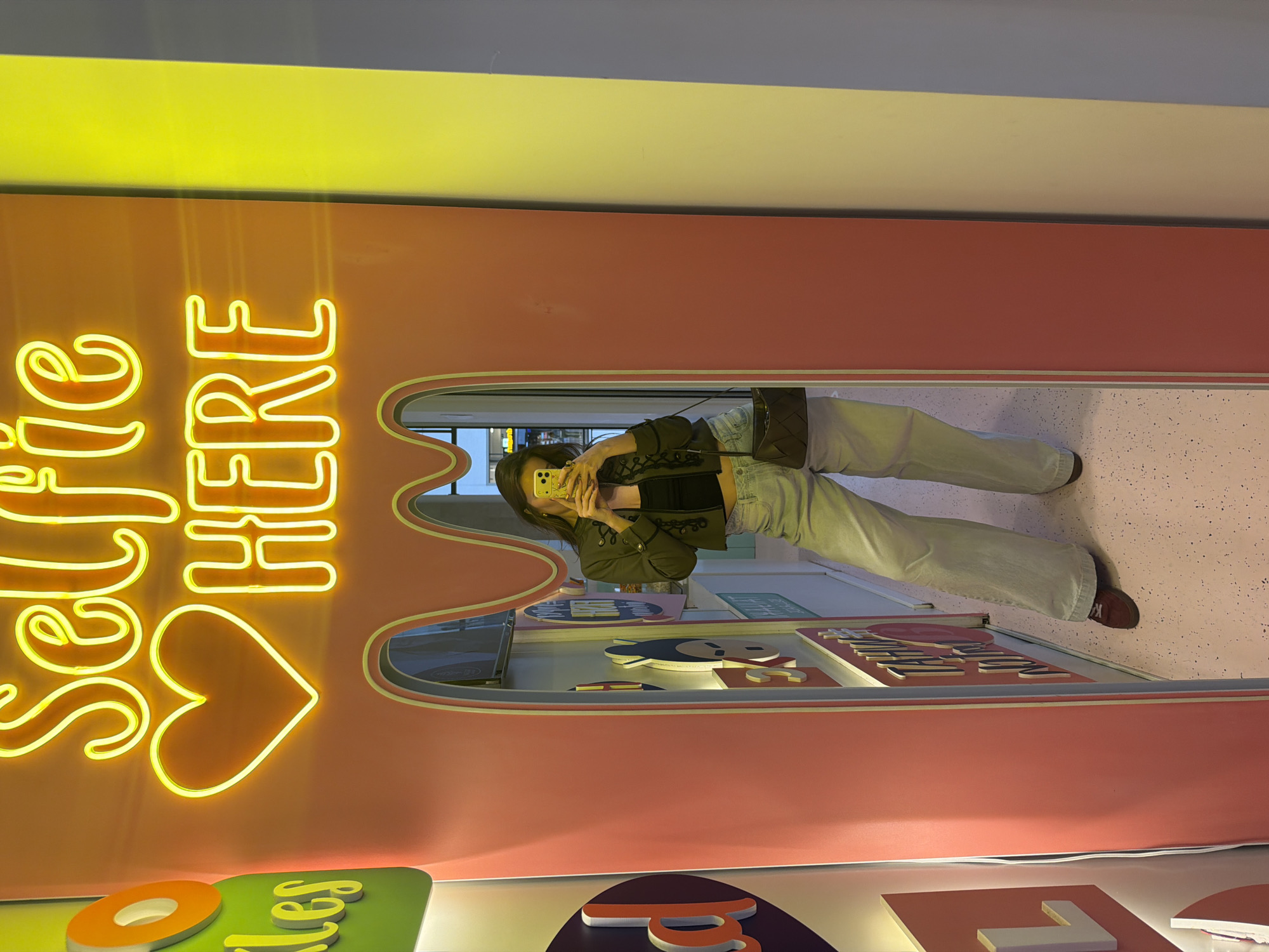 A person is captured in a mirror selfie within a vibrant, artificially lit indoor setting, likely in Belgrade, Serbia. The individual, dressed in a dark jacket over a light crop top, light blue wide-leg jeans, and brown shoes with a visible "K" on one, holds a yellow-cased smartphone to take the picture. A dark, woven shoulder bag hangs from their arm.
The backdrop features a prominent peach-pink wall with an eye-catching yellow neon sign that spells "Selfie HERE" and includes a heart symbol. Above this section, the wall is a solid yellow band, transitioning to a grey ceiling. The mirror itself is framed by an undulating peach-pink border. The floor is light-colored and speckled. Below the mirror, other colorful, indistinct graphical elements and displays are visible, suggesting the location is an interactive museum, a themed retail space, or a dedicated photo-op area. The overall aesthetic is modern and playful, designed to encourage photography.