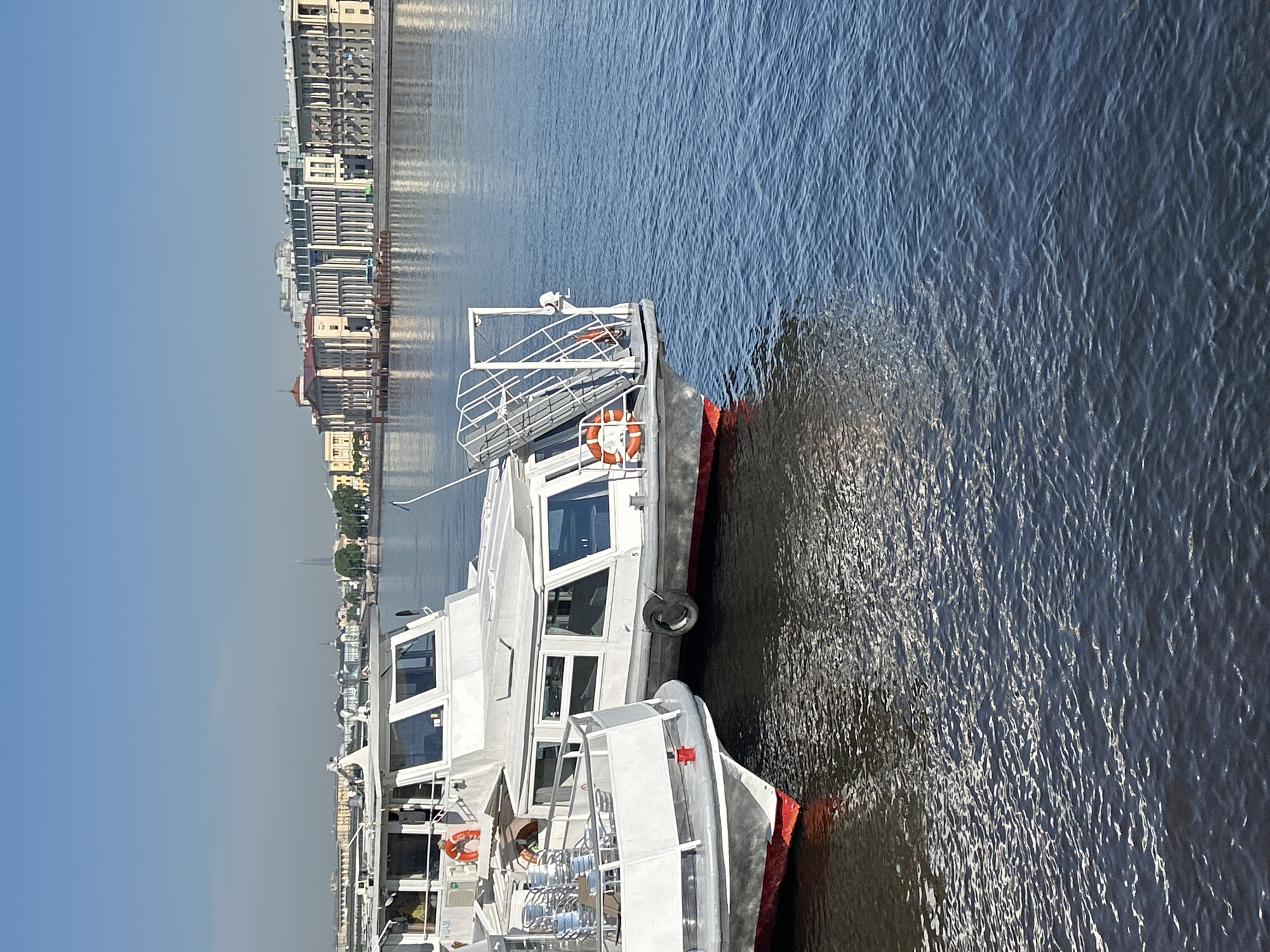The image depicts a bright, sunny scene along a wide body of water, likely a river or canal, flanked by a city skyline. In the foreground, two white passenger boats are prominently featured, appearing to be docked or moored side-by-side.
The boats are primarily white with red trim along their lower hulls. They possess multiple large windows, white metal railings on their decks, and a visible orange and white life preserver on each. Black rubber tires are used as fenders along the sides of the boats. One of the boats has the number "5" visible on its side.
The water surface is textured with numerous ripples and shimmering reflections, indicating strong sunlight. The color of the water is a deep blue-grey. Across the water, a dense city skyline stretches along the bank. The buildings exhibit a mix of architectural styles, from older, classical designs with distinctive roofs to more modern structures. The sky above the city is clear and light blue. In the far distance, a tall, slender, modern spire is faintly visible, potentially identifying the location.
The scene suggests a leisurely day in a bustling city known for its waterways, with the boats likely used for tours or public transport. No people are distinctly visible.