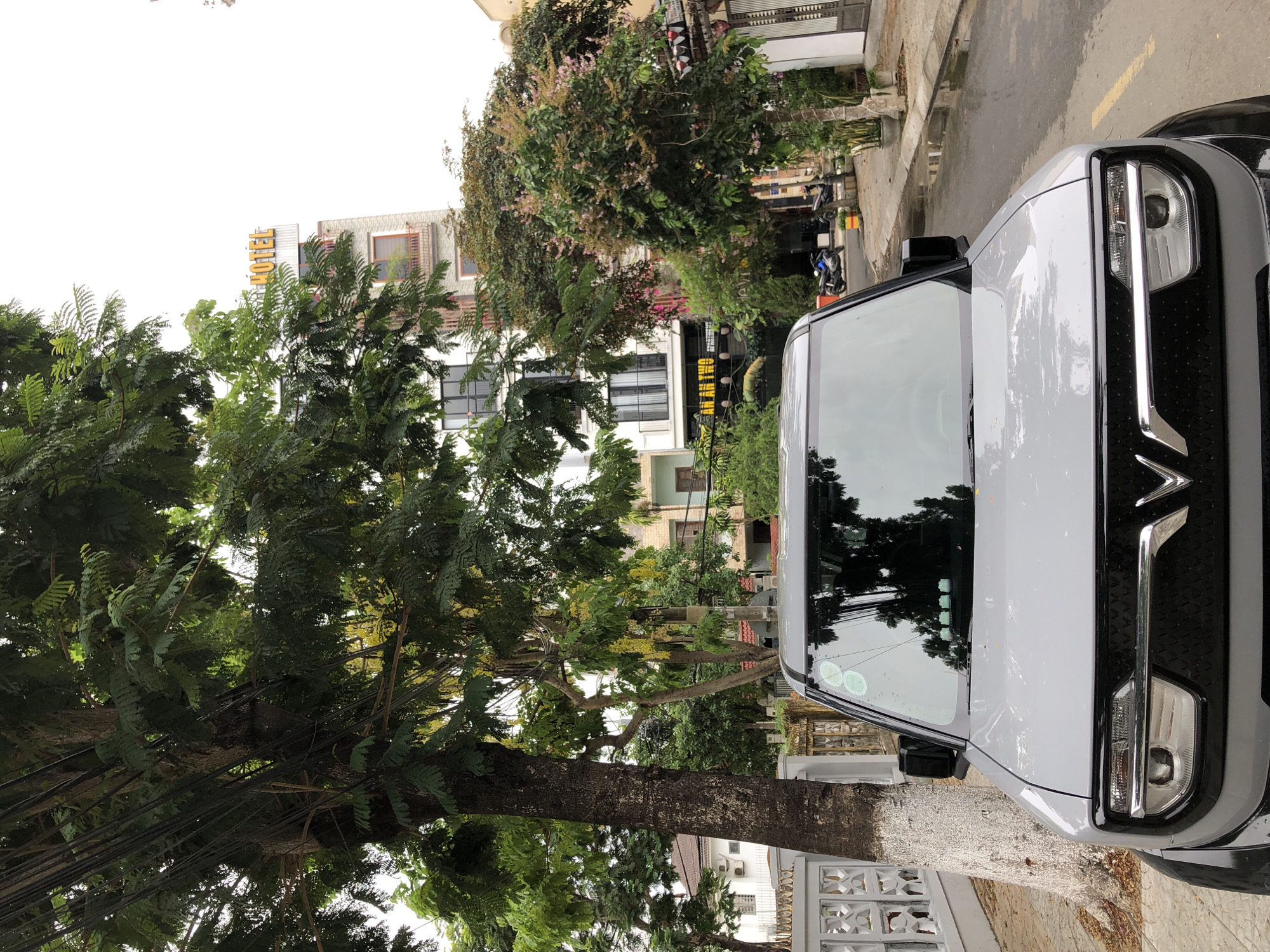 The image shows the front of a light gray electric car parked on a street in Da Nang, Vietnam. The car has a black grille with a V-shaped emblem and distinctive headlights. In the background, there is a tall building with a sign that reads "HOTEL" in yellow letters. There are also trees, other buildings, motorcycles, and a sidewalk with potted plants. The street appears wet, possibly from recent rain. The overall scene is urban and appears to be during the daytime.