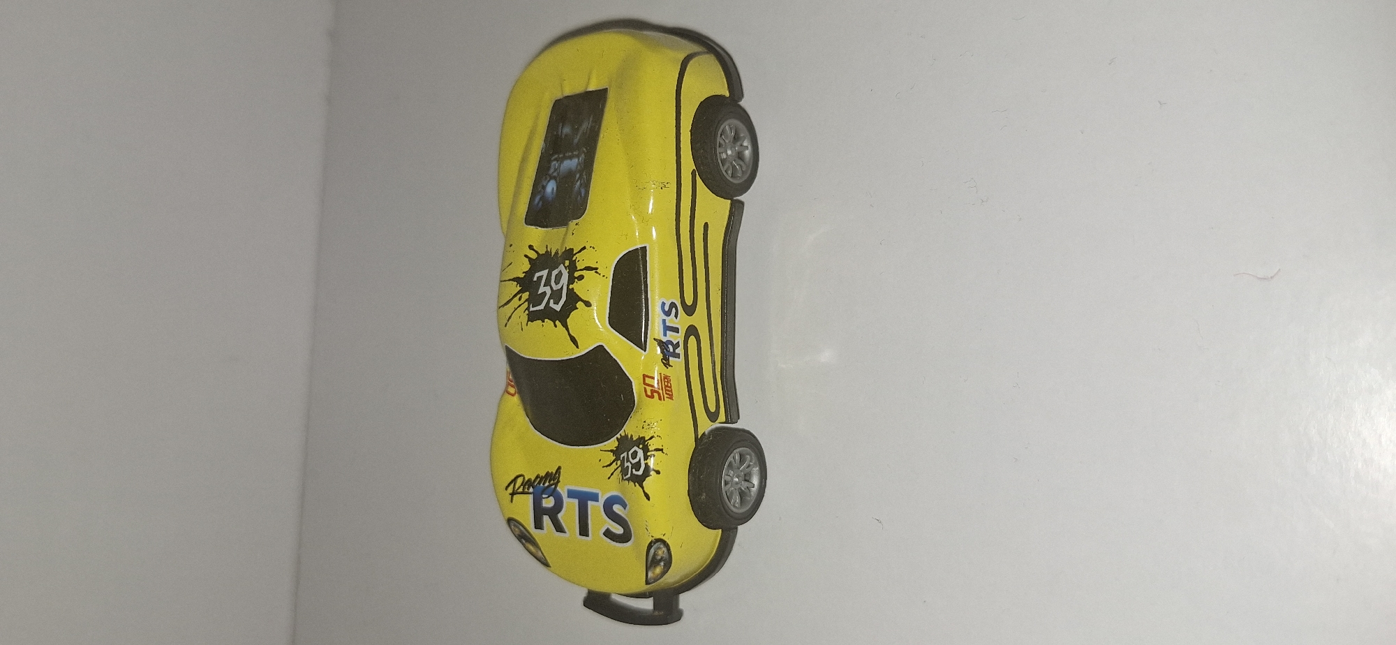 A yellow toy race car is displayed on a plain white surface, casting a subtle shadow. The car features black decals, including the number "39" within a splattered ink design, and "RTS" lettering in blue. The word "Racing" is also visible near the front of the car. The car has silver-colored wheels and a black spoiler at the rear. The image does not contain any identifiable landmarks or features that would allow for its location to be determined within Bila Tserkva, Ukraine.