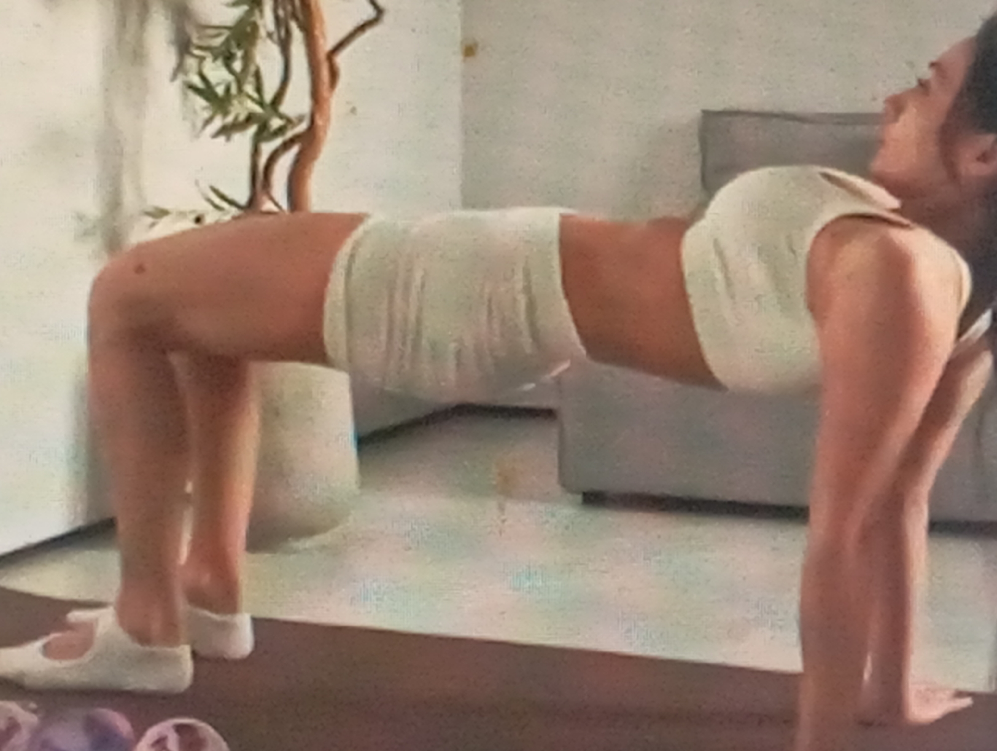 The image shows a woman in a white athletic outfit performing a crab walk exercise. She is on all fours with her hips lifted, forming an inverted U-shape with her body. Her hands and feet are on the floor, and her body is parallel to the ground. She is looking to the right, with a focused expression. The setting appears to be indoors, possibly a home gym or living room, with a light-colored wall and a plant in the background. A piece of furniture, likely a sofa, is also visible in the background. The lighting suggests it is daytime. There is no visible text in the image. The overall mood is one of exercise and focus.