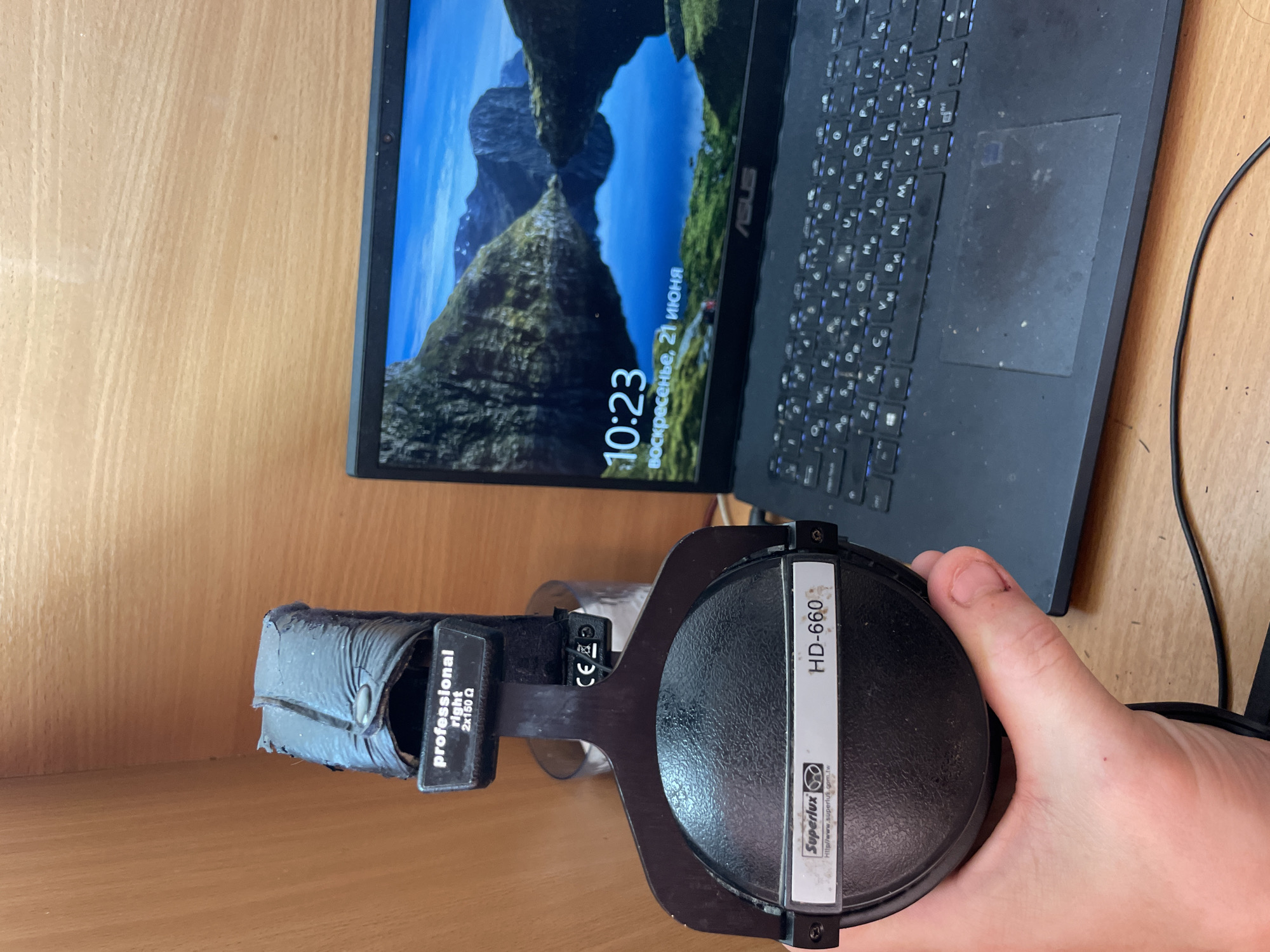 A pair of black Superlux HD-660 headphones, held by a person's left hand, is positioned in the foreground. The headphones have a "professional right 2x150Ω" label on them. To the right of the headphones, a black ASUS laptop is open, displaying a lock screen with a lake and mountain landscape. The time "10:23" and the date "воскресенье, 21 июня" (Sunday, June 21) are visible on the laptop screen. The background consists of a light-colored wooden surface, likely a desk or shelf, with a partial view of a glass container behind the headphones. The scene appears to be indoors, with artificial lighting. The image does not contain information about the city of Laishevo, Russia.