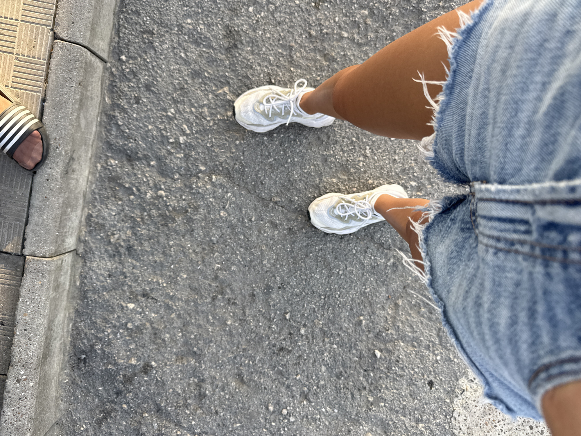 An overhead view shows a person's legs and feet as they stand on an asphalt surface next to a concrete curb. The person is wearing distressed denim shorts and white athletic shoes with laces. The curb is adjacent to a paved walkway. The scene is located in Cimișlia, Moldova.