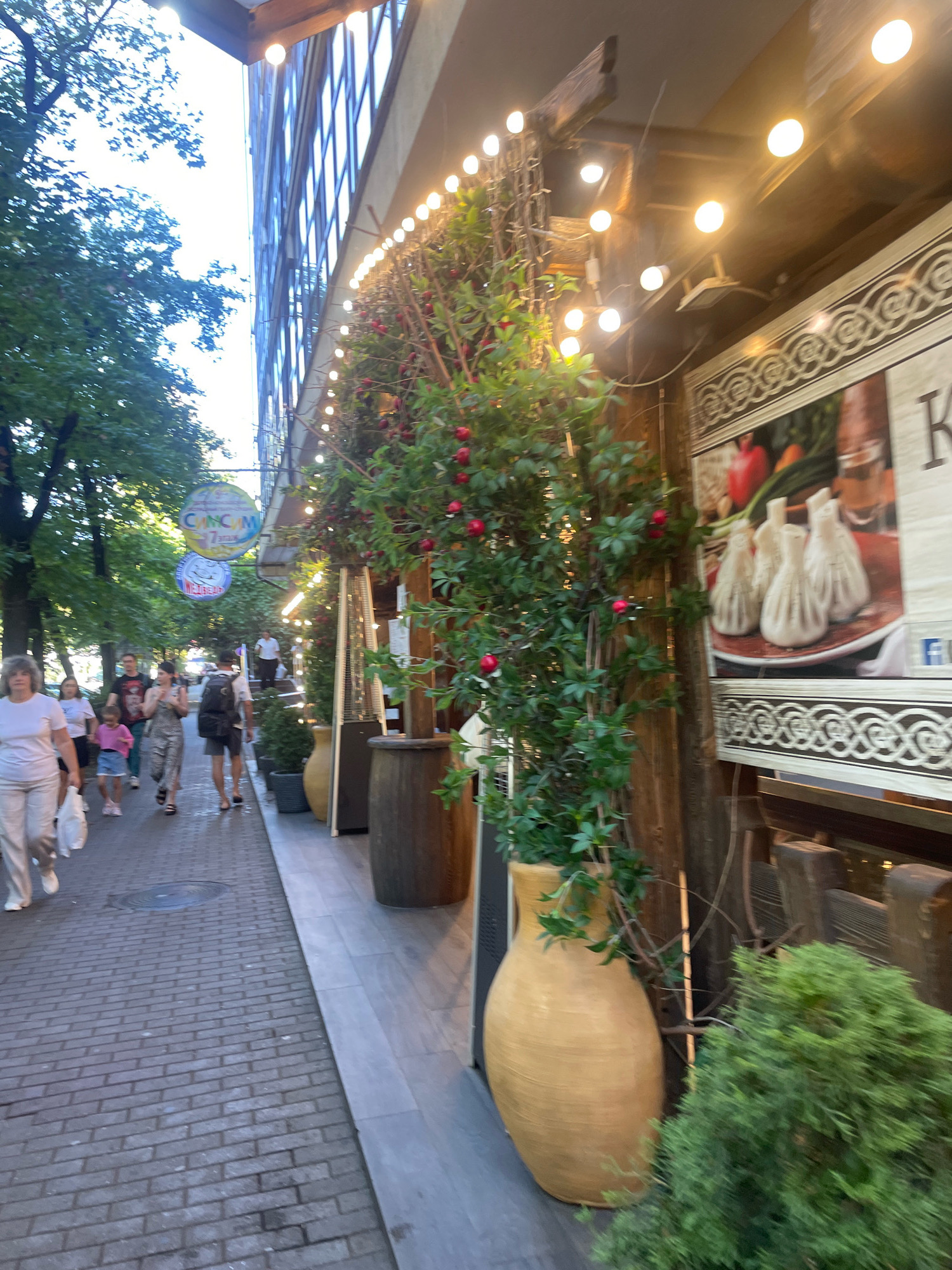 The image shows a street scene in Sochi, Russia, likely a sidewalk cafe or restaurant area. The left side of the image features a brick sidewalk lined with trees and a building with multiple windows. Several people are walking on the sidewalk. The right side of the image shows the facade of a building decorated with string lights and potted plants with red berries. A large poster displaying khinkali, a Georgian dumpling, is visible. A large, light-colored ceramic vase is in the foreground. The overall impression is of a vibrant outdoor dining or shopping area.