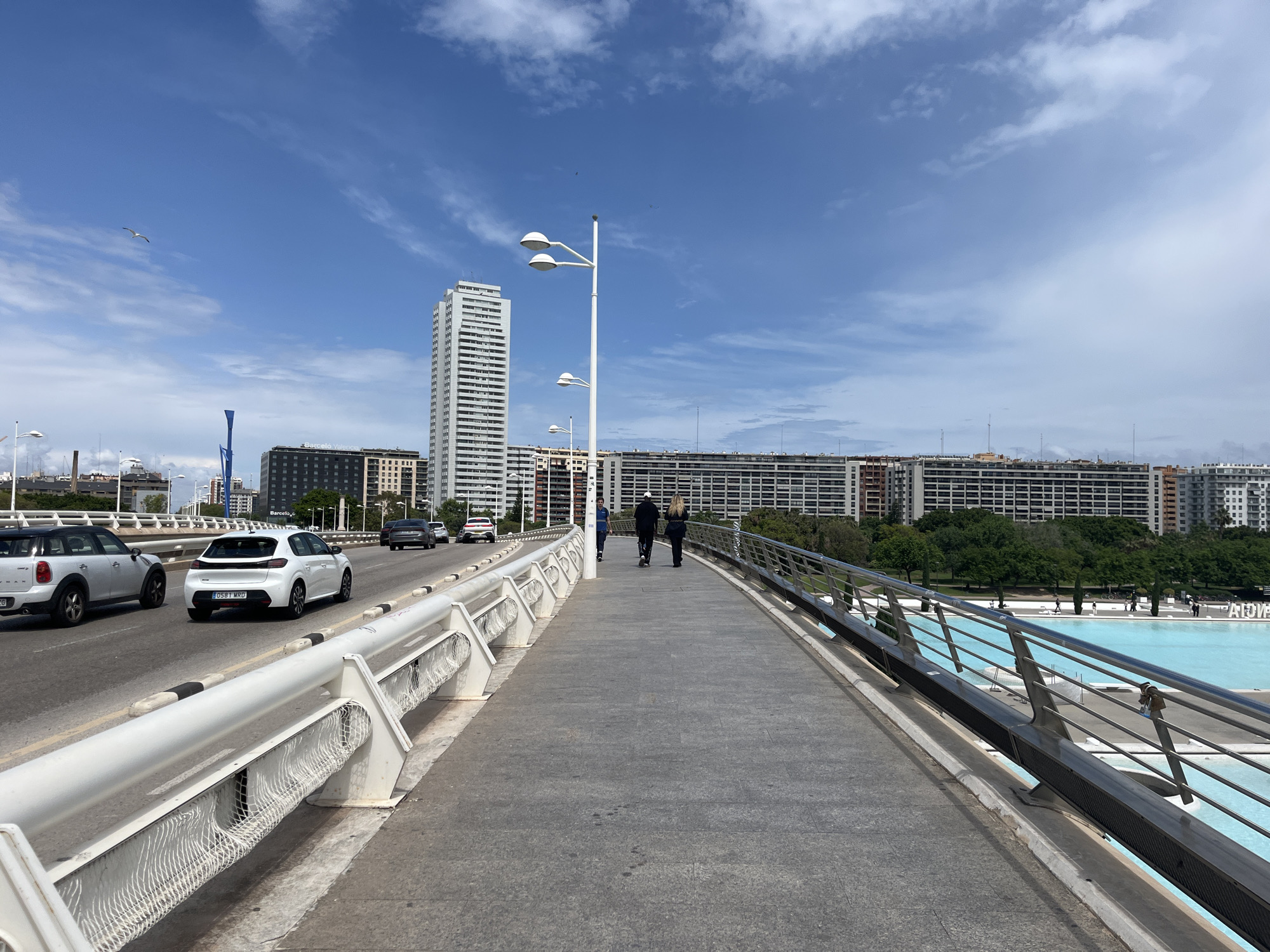 The image depicts a pedestrian perspective on the Puente de l'Assut de l'Or in València, Spain, on a bright, partially cloudy day. The bridge carries vehicular traffic on the left, with a white Mini Cooper and a white Peugeot visible. The pedestrian walkway occupies the center, with two individuals walking away from the camera. A taller man in dark clothing is followed by a taller woman. On the right side of the bridge, a park can be seen, and buildings are beyond the park. A reflective pool with blue water is below the bridge, and the walkway has steel railings. A tall, white skyscraper is visible in the background, flanked by other multi-story buildings. White lamp posts line the bridge's central walkway.