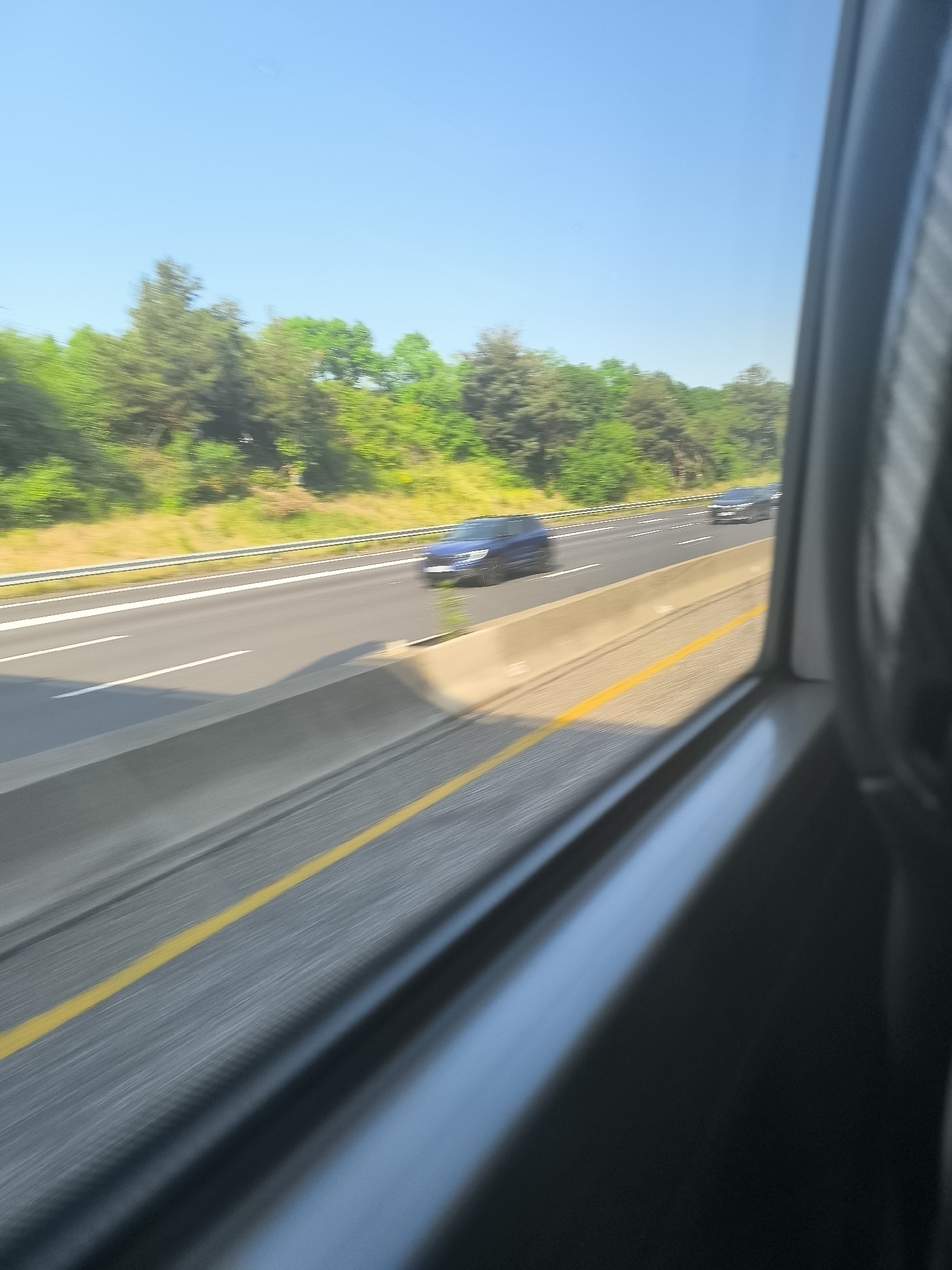 The image, likely taken from a moving vehicle, captures a view of a highway in L'Horme, France. The scene is dominated by the road stretching into the distance, with multiple lanes visible. A blue car is visible on the highway, and the tree line can be seen in the background. A yellow line is painted on the road. The window frame of the vehicle from which the photo was taken is present.