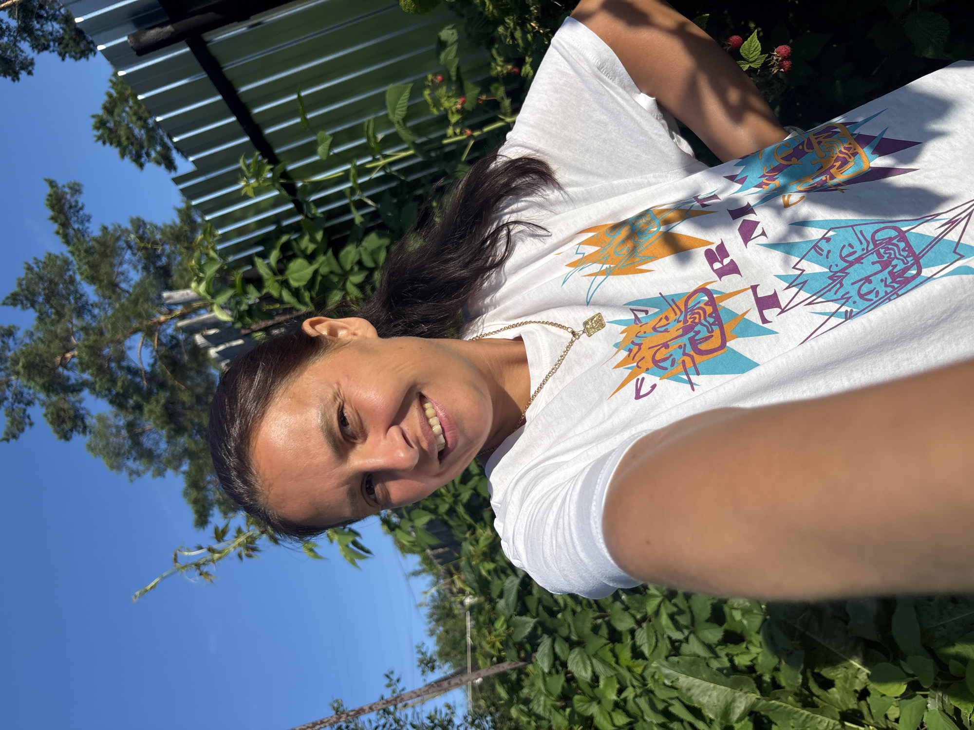 A woman takes a selfie in a garden, with lush green foliage and a metal fence in the background. The sky is clear and blue. The woman is wearing a white t-shirt with a colorful graphic print and a gold chain necklace. She is smiling and has her right hand on her hip. The image is a candid shot, likely taken outdoors on a sunny day, possibly in the city of Chaikovsky, Russia.