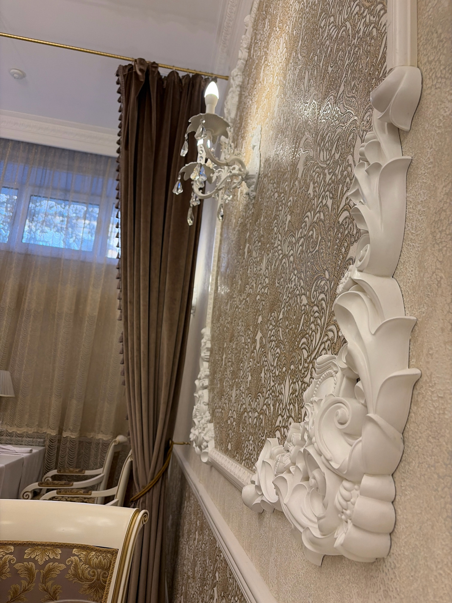 The image depicts an interior scene with ornate decorations. A textured wallpaper with a gold damask pattern covers the wall. Prominent white decorative molding, possibly plasterwork or stucco, is affixed to the wall, featuring intricate floral and scroll designs. A crystal sconce with a single light bulb is mounted on the wall to the left of the molding. In the background, rich brown velvet curtains are drawn back, revealing a window with sheer white curtains. To the left, a glimpse of a dining table set with a white tablecloth and chairs suggests a formal dining area. The furniture also appears to be upholstered in a patterned fabric with gold accents. The scene is likely from a residential or hospitality establishment in Angarsk, Russia, characterized by its opulent interior design. There are no people visible in the image.