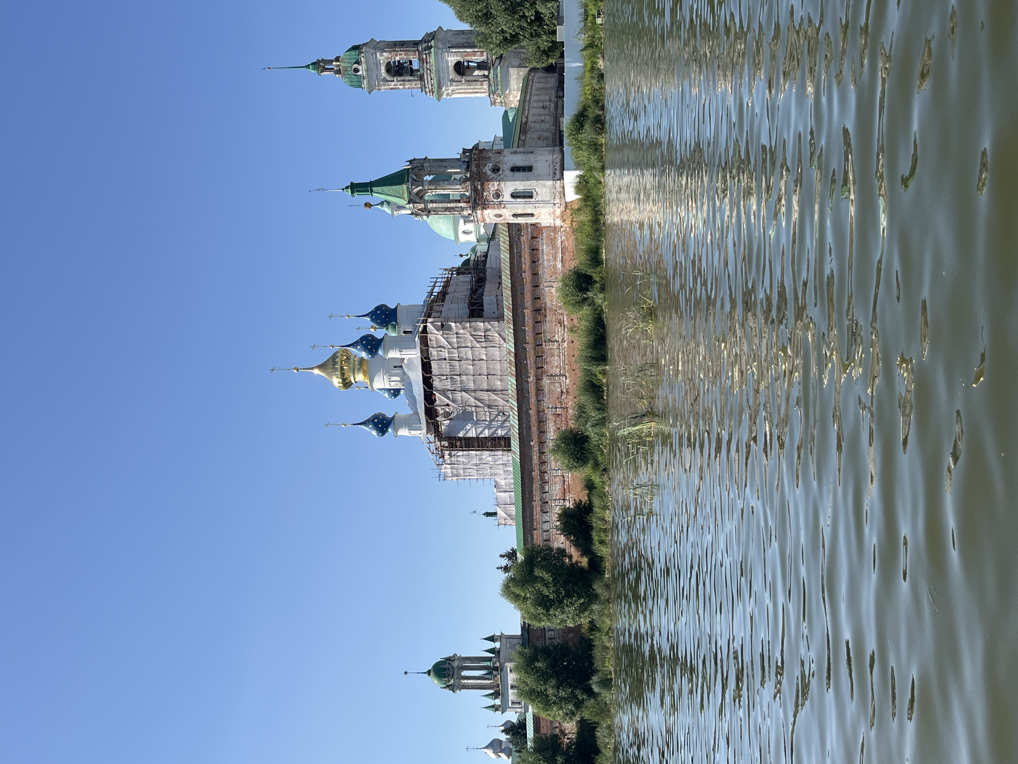 This is a wide, eye-level, outdoor, daytime, photographic, landscape shot of a monastic complex situated on the bank of a body of water. The sky is clear and blue. In the foreground, the water reflects the sky and the monastic complex, with ripples distorting the reflection. The monastic complex itself features several buildings with distinctive onion domes, some of which are gold, blue, or green. One of the larger buildings is partially covered in scaffolding and white sheeting, suggesting ongoing renovation or construction. A bell tower with a green roof and arched openings stands prominently to the right of the main building complex. A red brick wall surrounds the complex. Trees and greenery are visible along the bank between the water and the wall. The overall scene conveys a sense of historical and architectural significance, set against a natural backdrop.