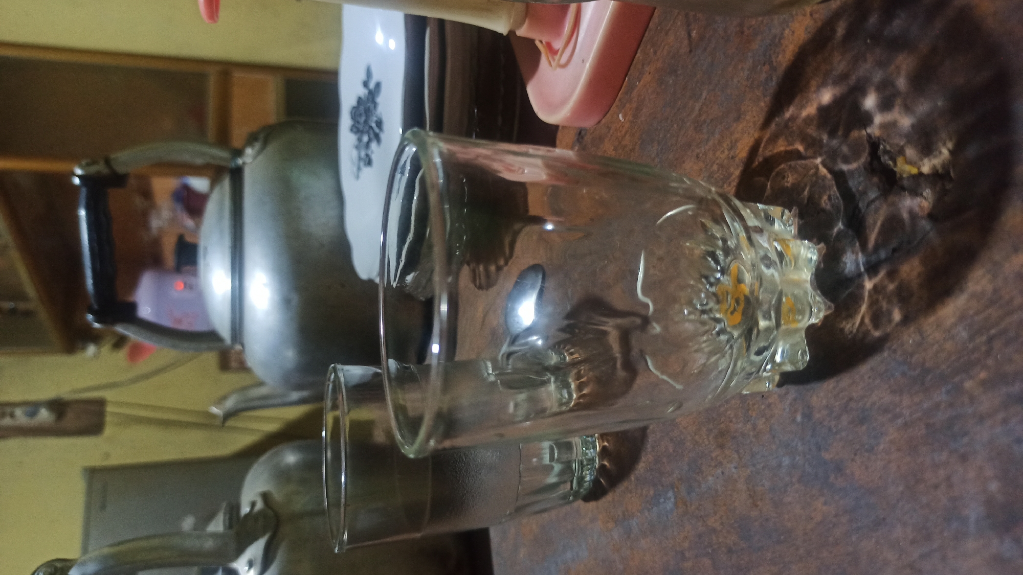 An indoor scene depicts two metal teapots, one larger than the other, placed on a wooden surface. In the foreground, two clear glasses are visible, one partially filled with a light-colored liquid and the other empty. The glasses are positioned on the same wooden surface. In the background, stacked plates with a dark floral pattern are present. A pink object, possibly a holder for something, is situated to the right of the plates. The lighting in the scene is dim, casting shadows on the wooden surface. The environment appears to be a domestic setting, consistent with a home in Tuba, Indonesia.