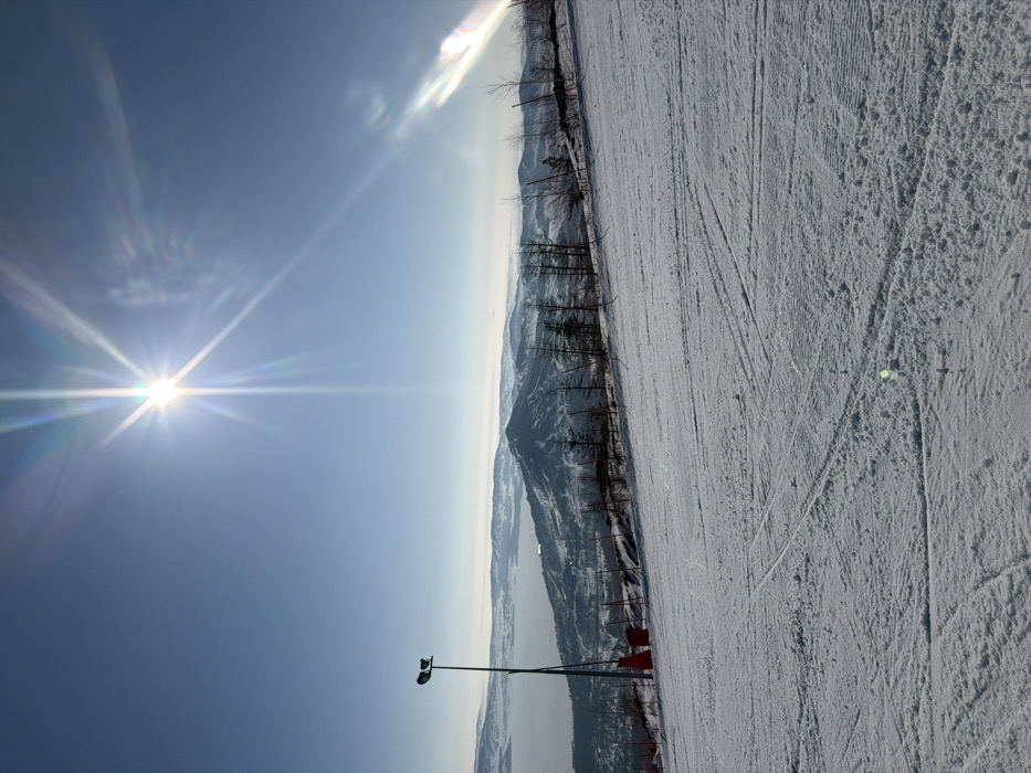 The image presents a wide view of a snow-covered ski slope in Zelenaya Polyana, Russia, under a clear, bright blue sky. The sun, located in the upper left quadrant, emits a prominent starburst lens flare with rainbow-colored diffraction spikes.
The foreground is dominated by white and light grey snow, exhibiting distinct ski tracks and areas of disturbed powder. In the midground, to the left, a tall, dark-colored pole with two downward-angled light fixtures is vertically positioned. Below this pole, red protective pads or markers are visible at the edge of the slope. Further to the right along the midground, sparse deciduous trees, mostly bare, and some small coniferous trees are scattered across the rising terrain, interspersed with red marker poles that delineate the slope's edge.
The background features multiple layers of snow-covered mountain ranges and hills receding into the distant horizon. A hazy body of water, appearing as a light grey expanse, is visible in the valley to the left, partially obscured by atmospheric conditions. The overall scene is brightly illuminated by direct sunlight. No individuals or animate subjects are present in the frame.