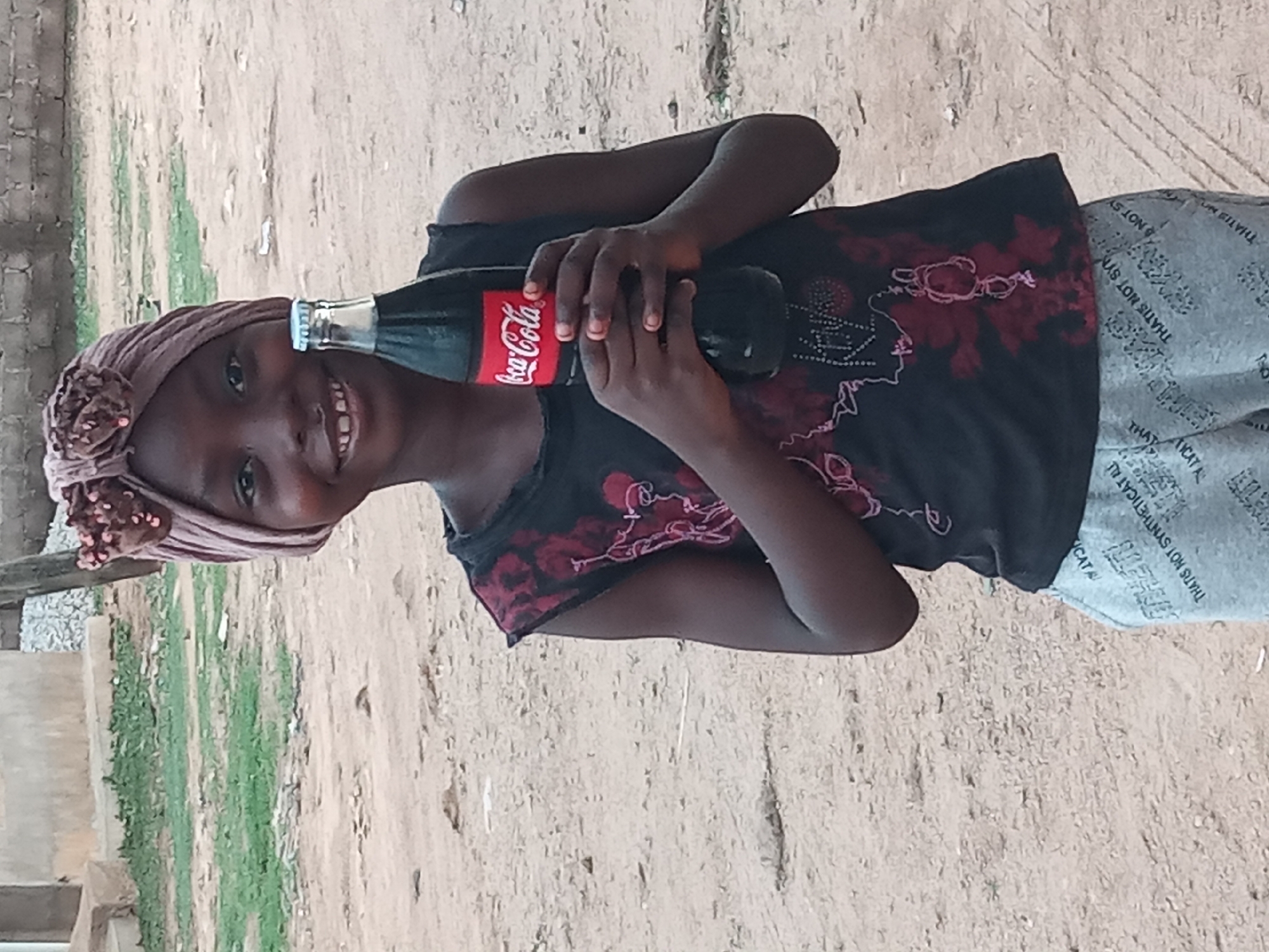 A young girl stands outdoors in Sabon Tasha, Nigeria, holding a glass bottle of Coca-Cola. She wears a headwrap, a dark-colored top with a floral pattern, and printed gray pants. The background shows a dirt and grassy area with a partial view of a building.