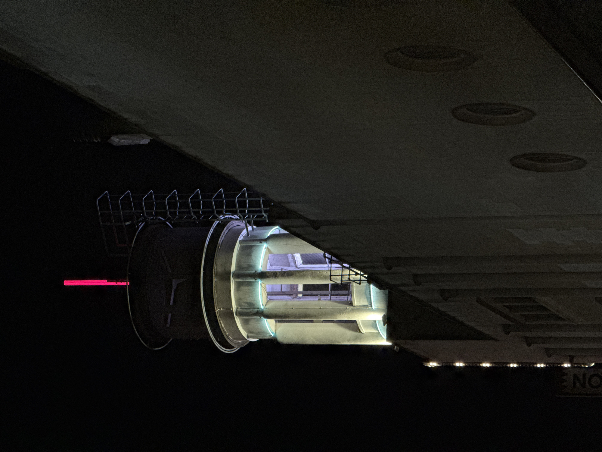 The image shows a low-angle, nighttime view of what appears to be a portion of a modern building, likely a structure within the city of London. The dominant feature is a cylindrical structure with a metallic frame, displaying internal illumination that gives off a pale blue light. To the left of this structure is a red horizontal light. Above the cylindrical structure is a large, flat, dark surface with evenly spaced circular indentations, possibly part of the building's architecture or a ceiling feature. The overall environment is dark, suggesting the image was taken at night, with minimal ambient lighting. The presence of the cylindrical structure and building elements gives an impression of modern architectural design.