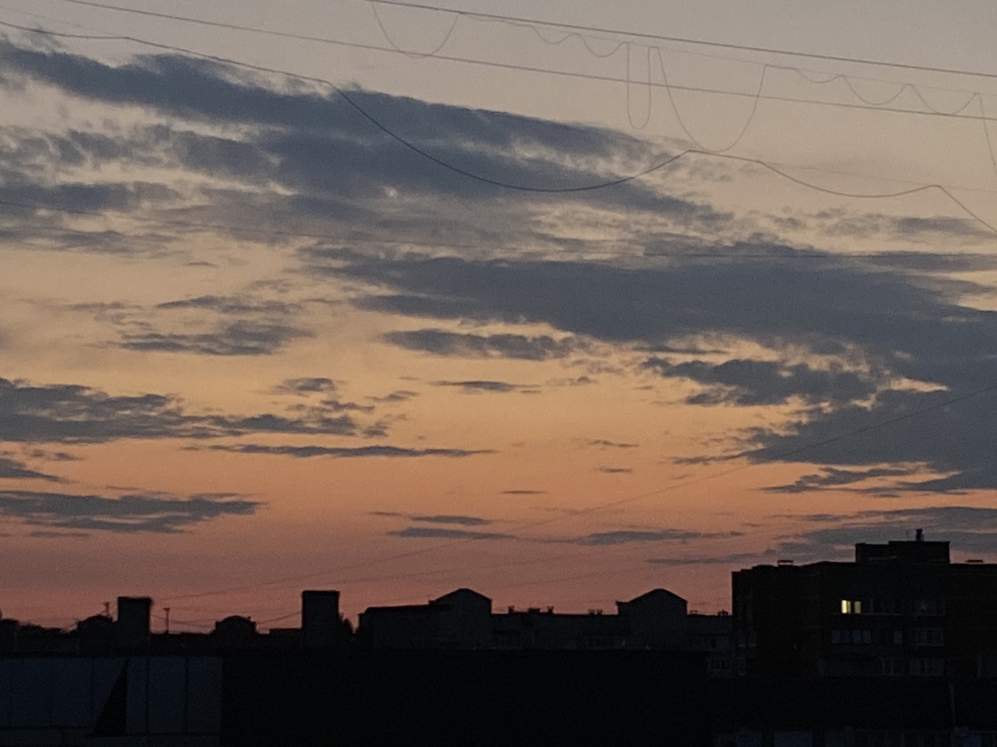 The image depicts a silhouetted cityscape against a dusky sky. The sky is a gradient of orange and pink hues, with scattered grey clouds. Several buildings, varying in height and architectural style, are visible in silhouette along the horizon. Some of the buildings have visible windows, with one building in the lower right corner showing two illuminated windows. Overhead, there are visible utility wires stretching across the frame. The overall scene suggests an urban environment at dusk or dawn.