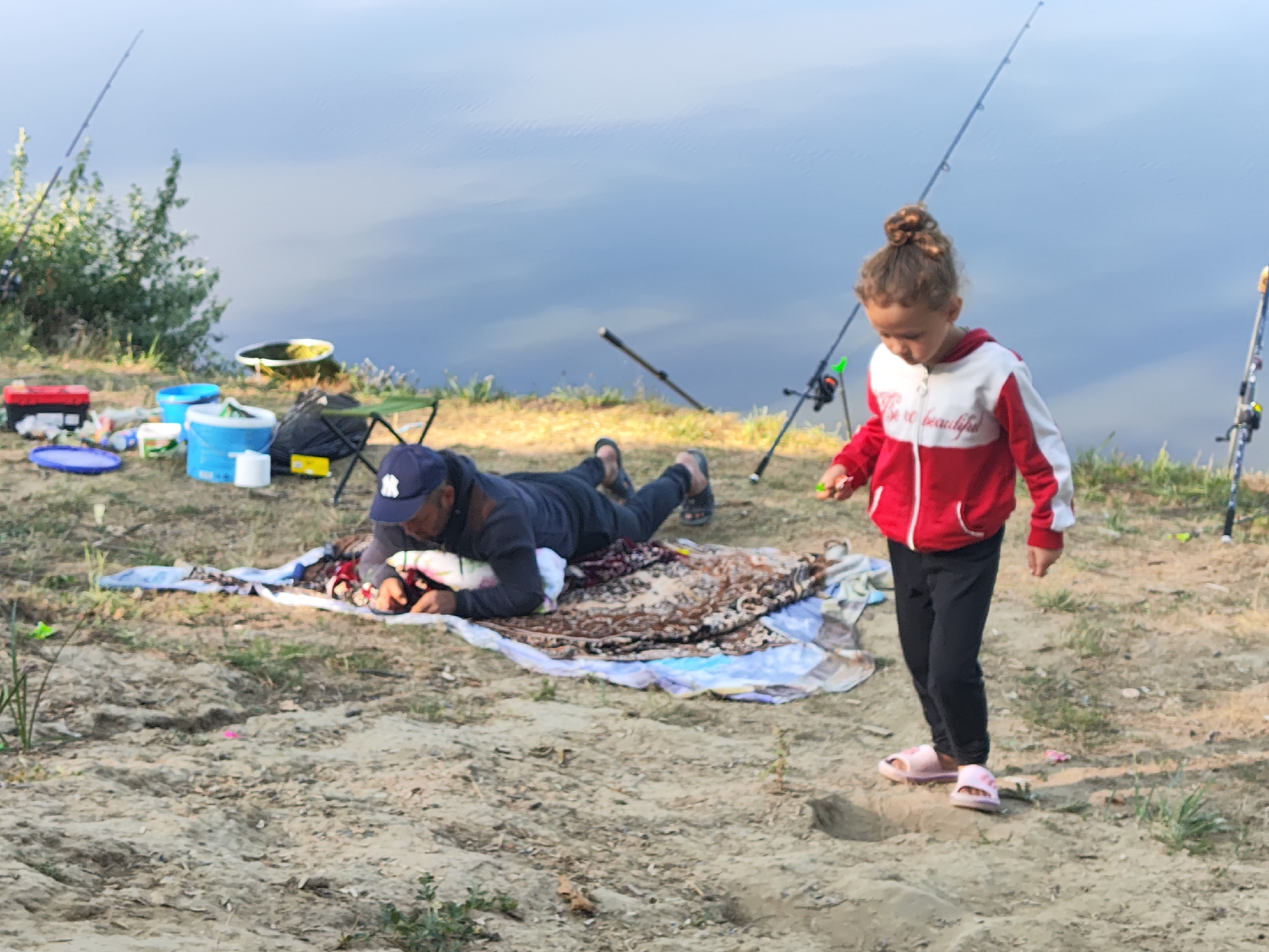 In Găleștii Noi, Moldova, on a sandy riverbank, a man in a dark blue cap and jacket lies on a patterned blanket next to fishing gear and several blue and white buckets. A young girl in a red and white hooded jacket and black pants stands nearby, facing away from the man, and appears to be holding something in her right hand. The river, with calm, reflective water, forms the background, with fishing rods extending into the scene from both the left and right. Green foliage is visible on the left side of the image, and sparse grass dots the sandy ground.