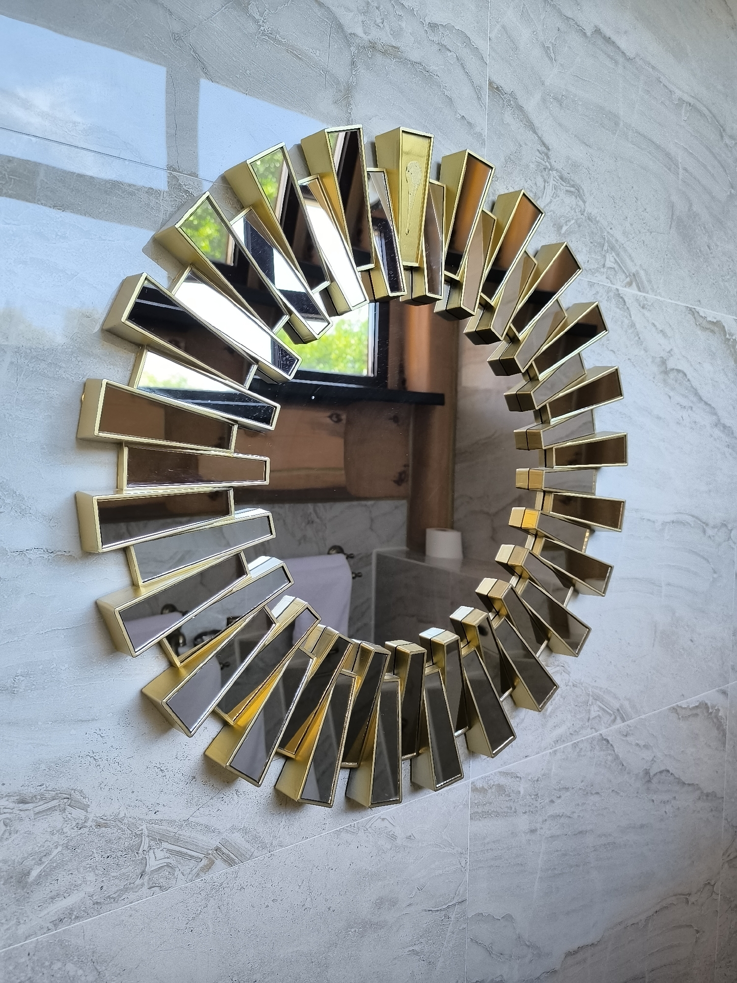 A sunburst mirror with a gold frame and beveled glass is mounted on a marble-tiled wall. The mirror reflects a room with wooden elements and a white roll of toilet paper. The wall tiles have a gray and white marbled pattern. The sunlight casts shadows on the wall.