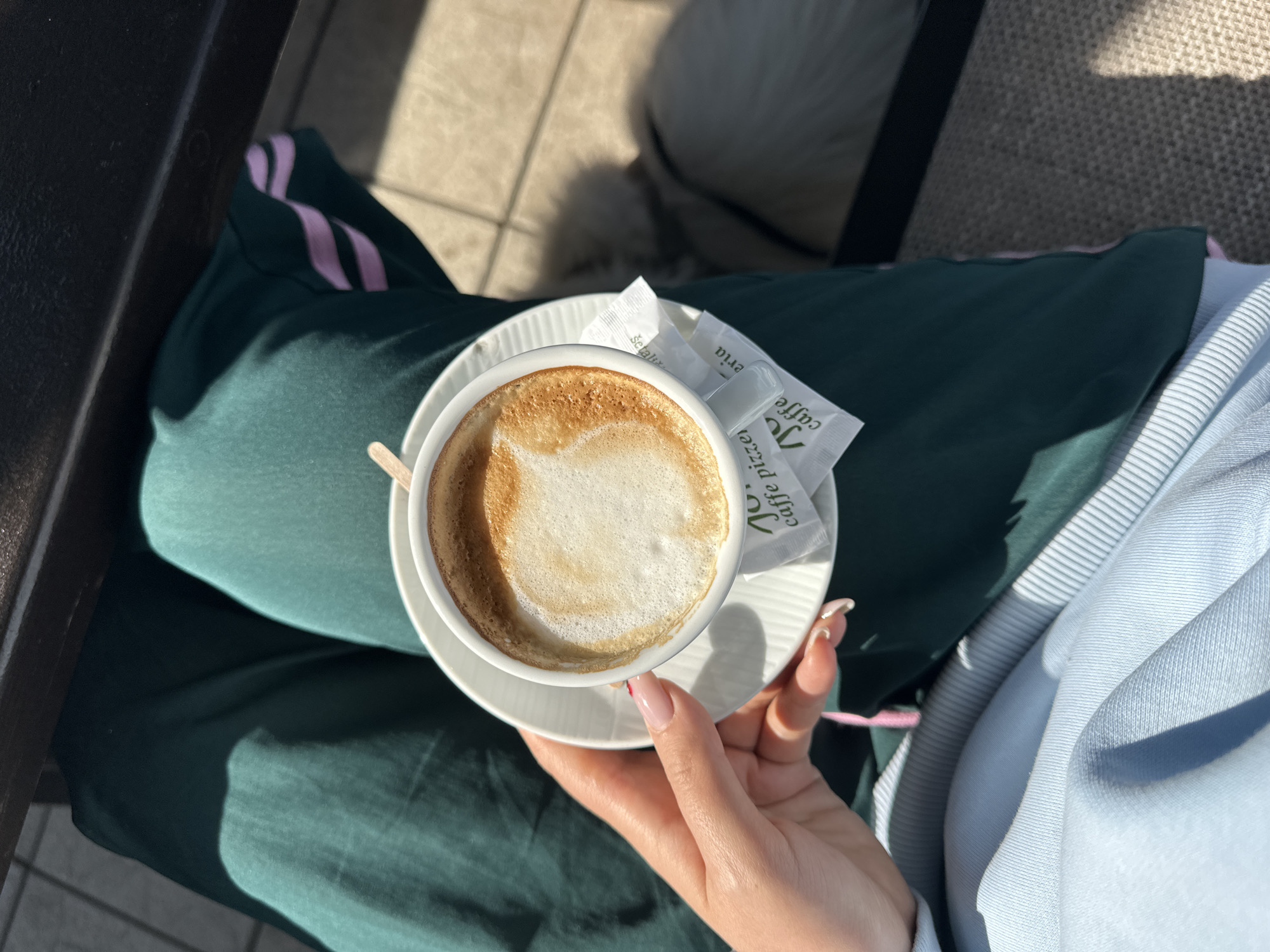A woman is holding a cup of cappuccino on a saucer in her right hand. Sugar packets sit beside the cup on the saucer. She is wearing green pants with pink stripes and a light blue hoodie. The background suggests an outdoor setting, likely a cafe or terrace in Šušanj, Montenegro. The specific location in Šušanj cannot be determined from the image.