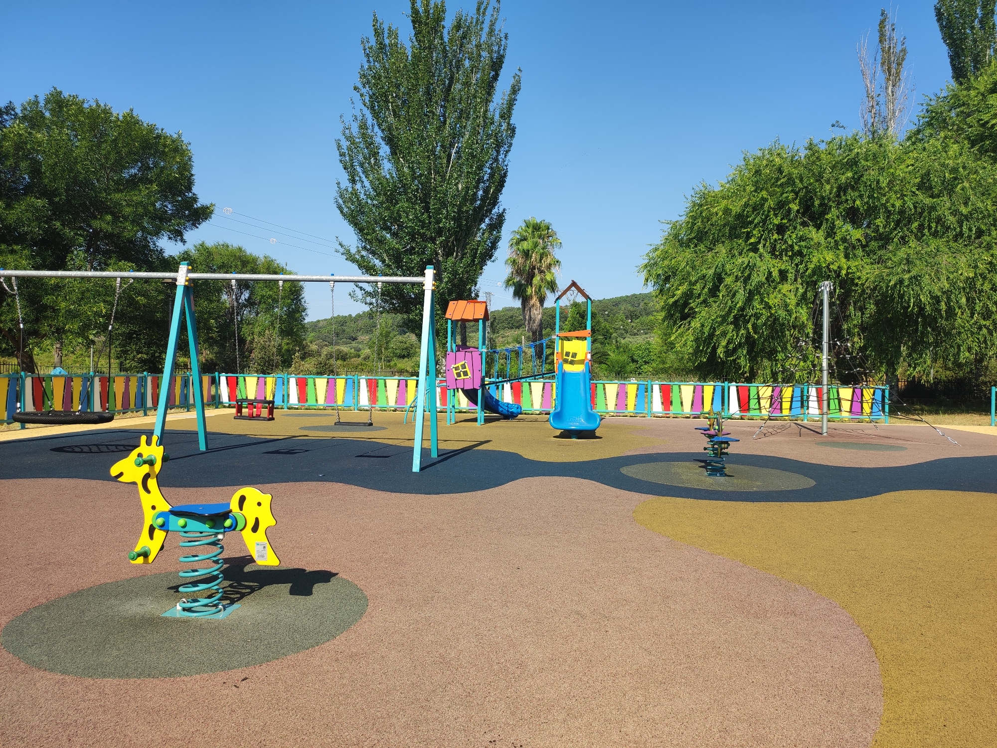 A bright, sunny day illuminates a vibrant children's playground located in Cadalso, Spain. The ground is a mix of soft, speckled surfaces in brown, dark grey, and yellow, artfully arranged in smooth, curving patterns. In the foreground, a yellow spring-mounted rocking toy shaped like a giraffe is centered on a grey circular mat. Behind the giraffe, a swing set with teal supports and multi-colored swings stands on a dark grey surface, bordered by a fence of alternating colored panels (pink, yellow, blue, green, orange, red). Further back, a colorful play structure features a red-roofed section with a yellow window, a blue slide, and a rope bridge connecting two parts. To the right of this structure is a small, multi-colored spring rider with a blue seat. The background is dominated by lush green trees and foliage, with a clear blue sky overhead. Power lines are visible strung between poles in the distance, against a gently sloping, tree-covered hillside. The overall impression is a clean, well-maintained, and inviting public play area.