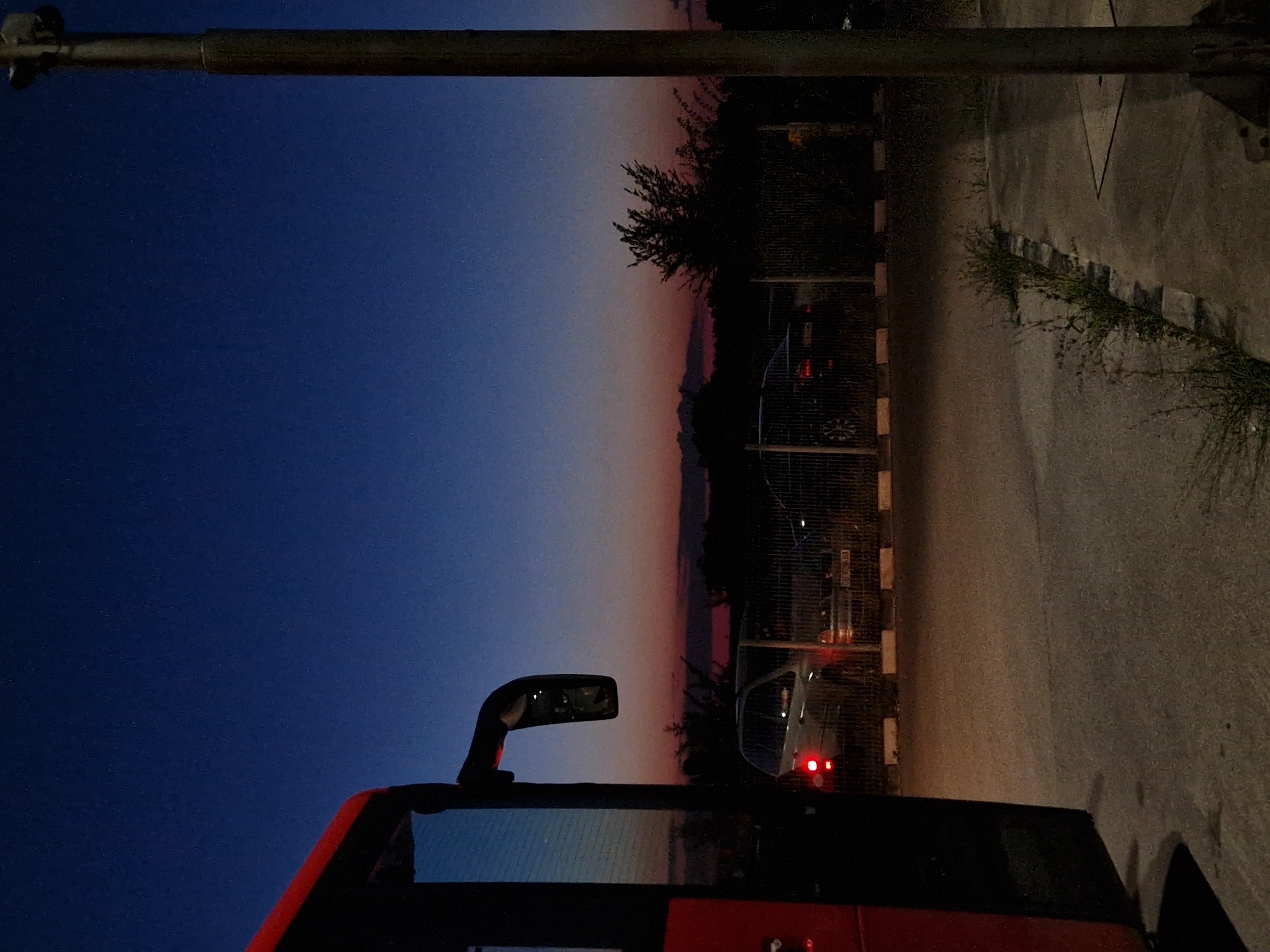 The image captures a roadside scene at dusk, with a large red bus occupying the left foreground. The bus's side is dark, with its reflection visible in the large side mirror mounted on the right side of its front. Behind the bus, a chain-link fence encloses a parking area. Two cars are parked within this area, their headlights and taillights illuminated. The car on the left appears to be a silver SUV, while the car on the right is darker. The ground is paved, with a curb separating the parking area from a slightly elevated sidewalk or walkway. Overgrown grass and weeds are visible along the edge of this walkway. In the background, a line of trees and foliage is silhouetted against the twilight sky, which transitions from a deep blue at the top to hues of pink and orange near the horizon. A tall, dark pole, possibly a lamppost, stands vertically on the right side of the frame. The overall atmosphere is quiet and transitional, characteristic of the hour between daylight and night. The location is identified as being in the city of Bahovitsa, Bulgaria.