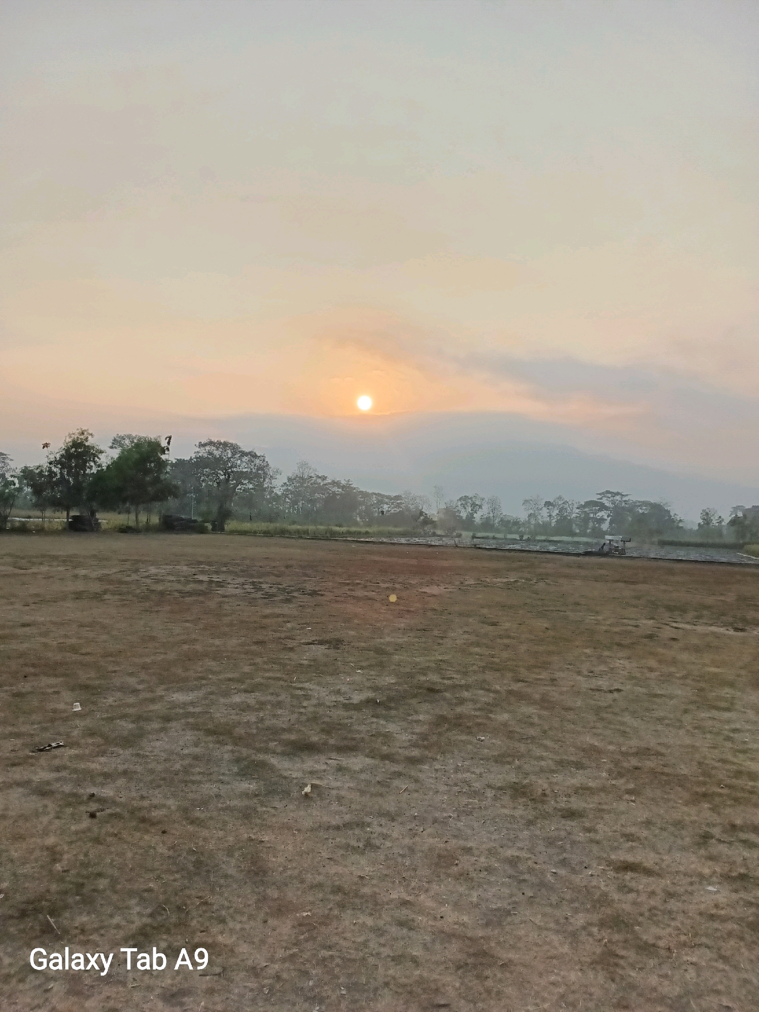 The image depicts an outdoor scene at what appears to be dawn or dusk, with the sun visible as a bright orb low in the sky, partially obscured by clouds. The landscape consists of a large, sparsely vegetated open field in the foreground, with a line of trees and distant hills forming the background. The ground is mostly dry and appears to be a mix of soil and short, dry grass. Several scattered objects, possibly litter, are present on the field. To the left of the frame, near the trees, there are a few indistinct structures or objects, and further into the scene, a small vehicle is visible. The sky is a pale orange and white, indicating diffused sunlight. The overall impression is one of a rural or undeveloped area. The watermark "Galaxy Tab A9" is present in the lower left corner of the image.