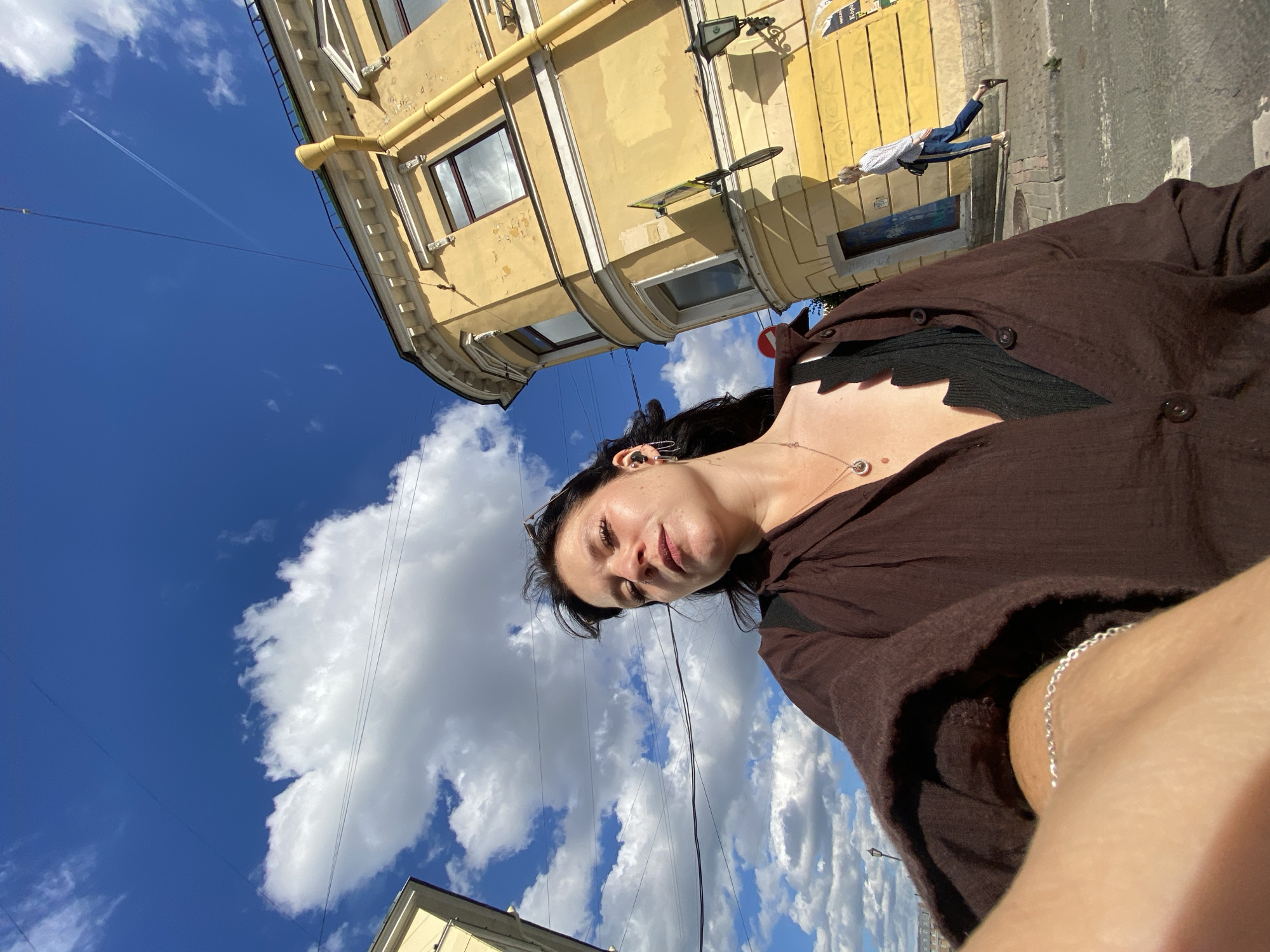 The image shows a woman taking a selfie outdoors in Saint Petersburg, Russia. She is wearing a brown button-up shirt over a black top and a necklace. Her arm, wearing a silver bracelet, is extended towards the camera. Behind her, a yellow building with ornate architectural details is visible. The sky is blue with scattered white clouds. A woman in a patterned shirt and jeans is walking in the background. The scene appears to be on a street with a sidewalk and road.