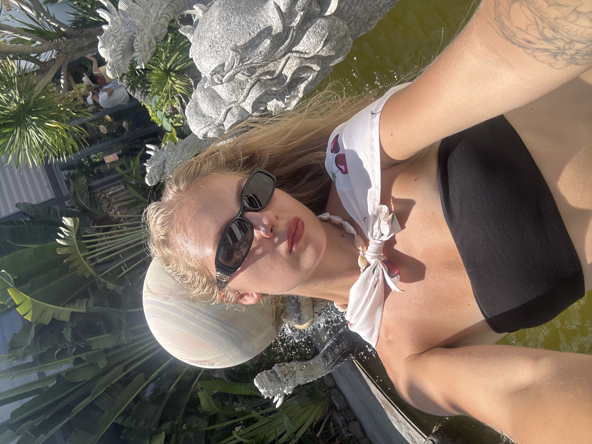 The media file shows a selfie of a young woman with blonde hair, wearing sunglasses and a white top with a cherry pattern tied around her neck. She is wearing a black bikini top. The scene appears to be outdoors, possibly near a pond or water feature, with lush green foliage in the background. There are also stone dragon statues visible. The lighting suggests it is daytime, with bright sunlight casting shadows. The woman's expression is relaxed and she appears to be enjoying herself. The location is identified as Tuy An Nam, Vietnam. There is a faint "GUCCI" logo visible on the side of her sunglasses.